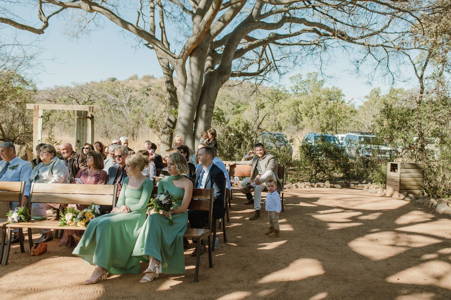 Wedding guests seated on rustic wooden benches under shady trees during an outdoor desert ceremony.