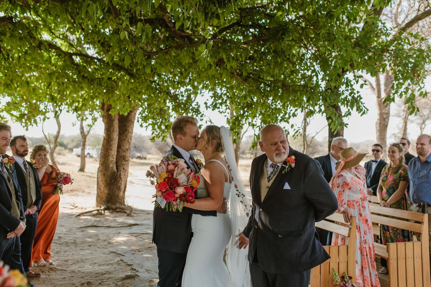 Wedding guests mingle under a canopy of green leaves during an outdoor reception.
