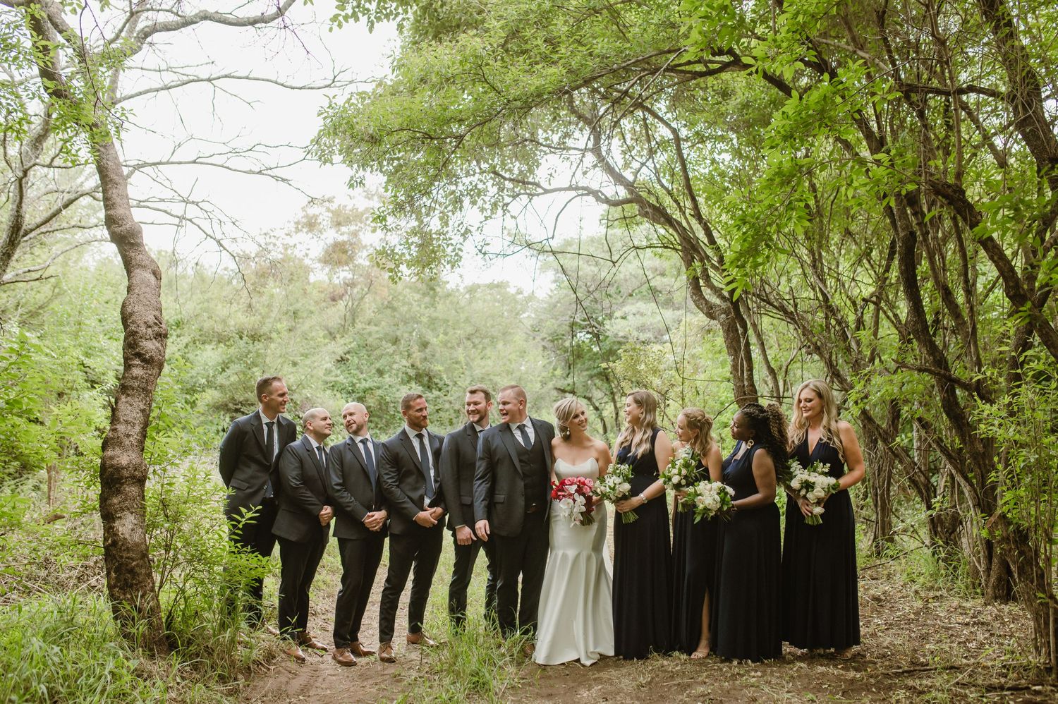 A wedding party poses for photos in a lush green wooded setting.