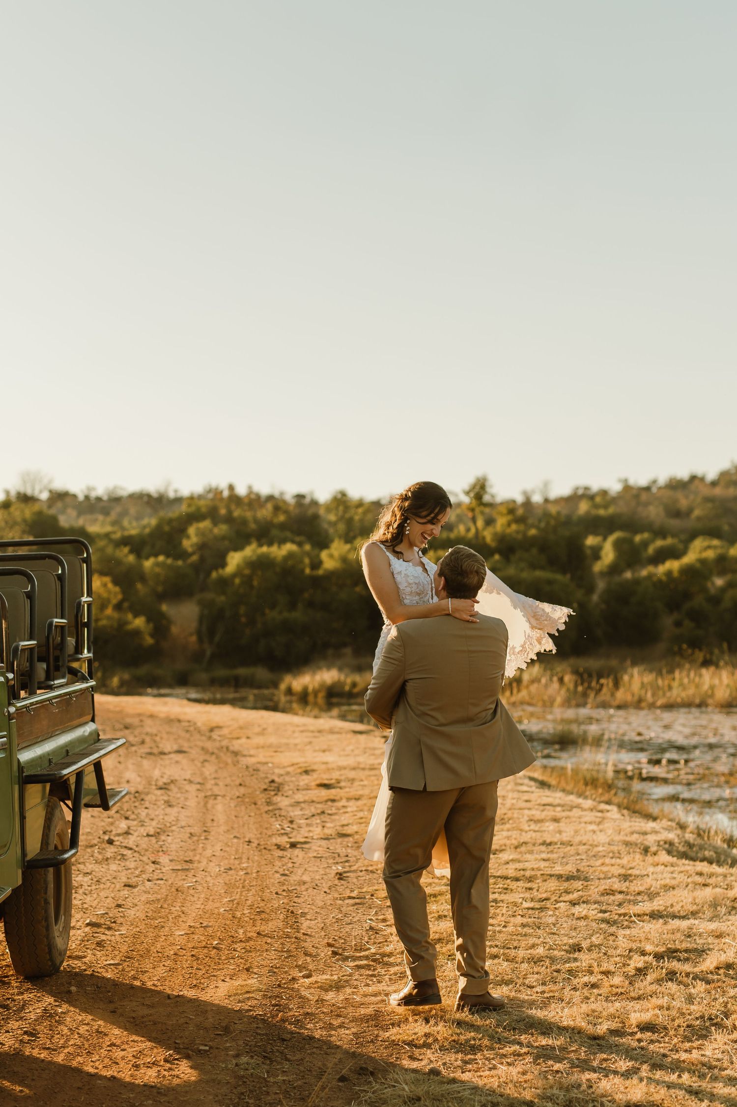 A couple embraces during golden hour on a dirt road with an off-road vehicle in the background.
