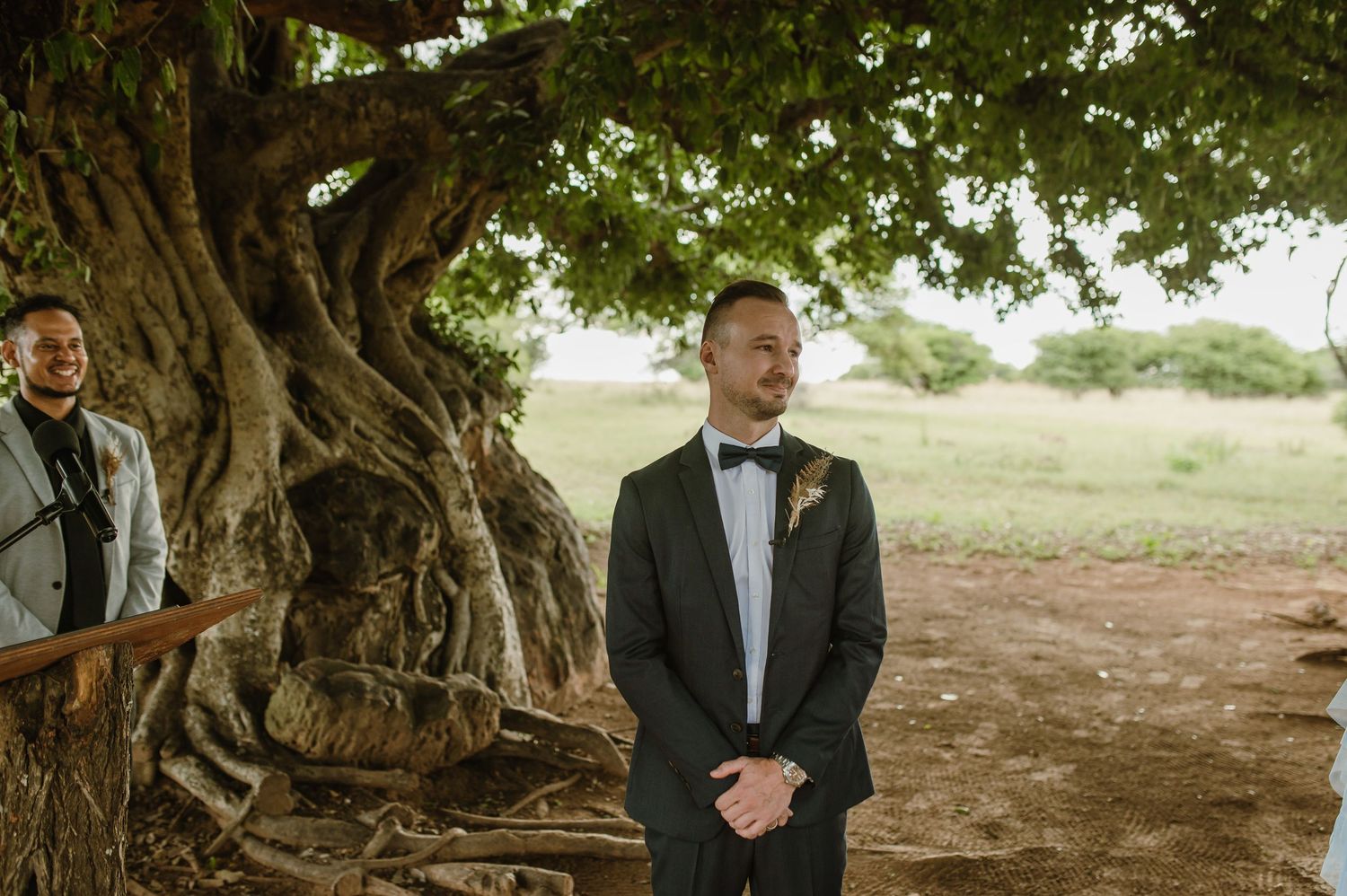 A groom in a black tuxedo stands waiting under a large tree at an outdoor wedding venue.