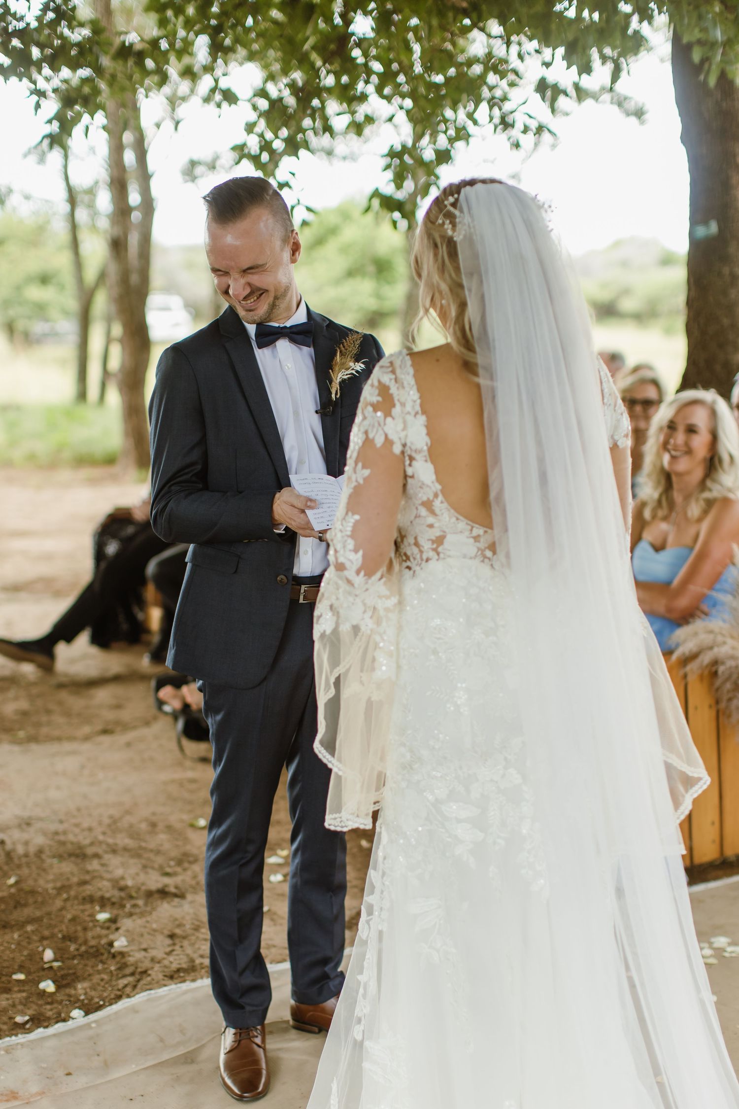 A couple shares a joyful moment during their outdoor wedding ceremony under a canopy of trees.