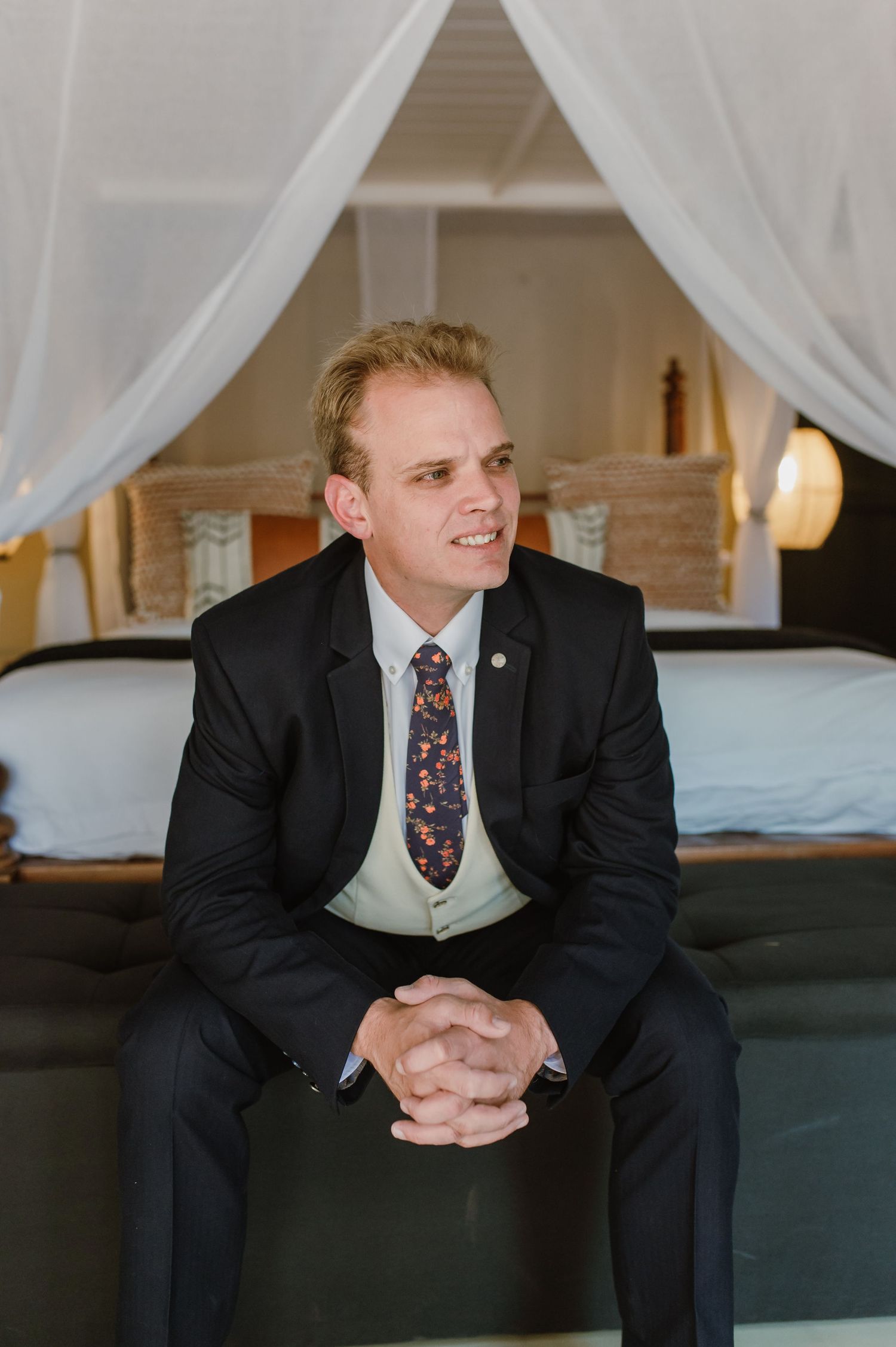 Person in formal suit and vest sits on dark couch in elegant bedroom setting.