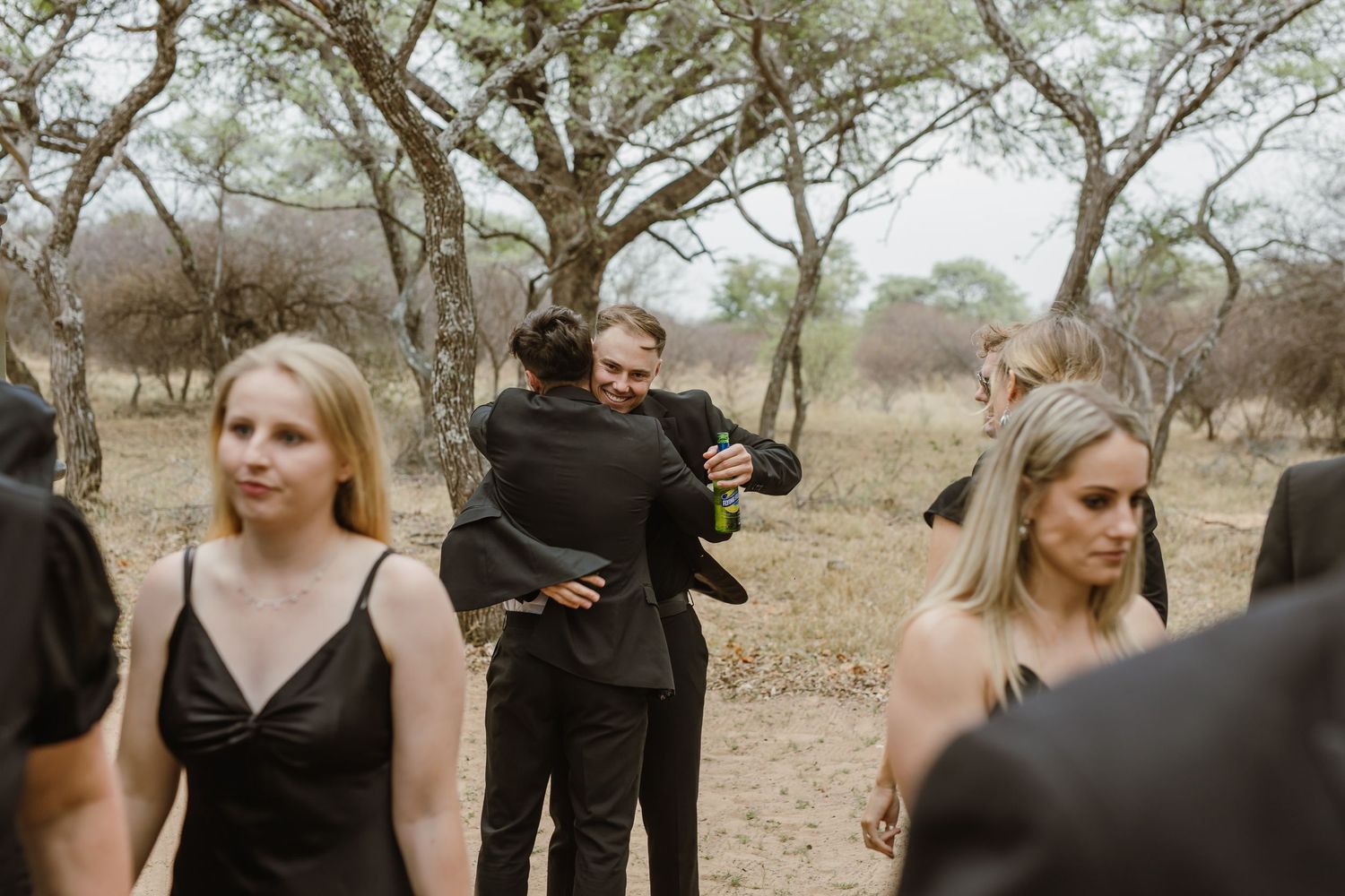Wedding guests in formal attire mingling outdoors among trees.