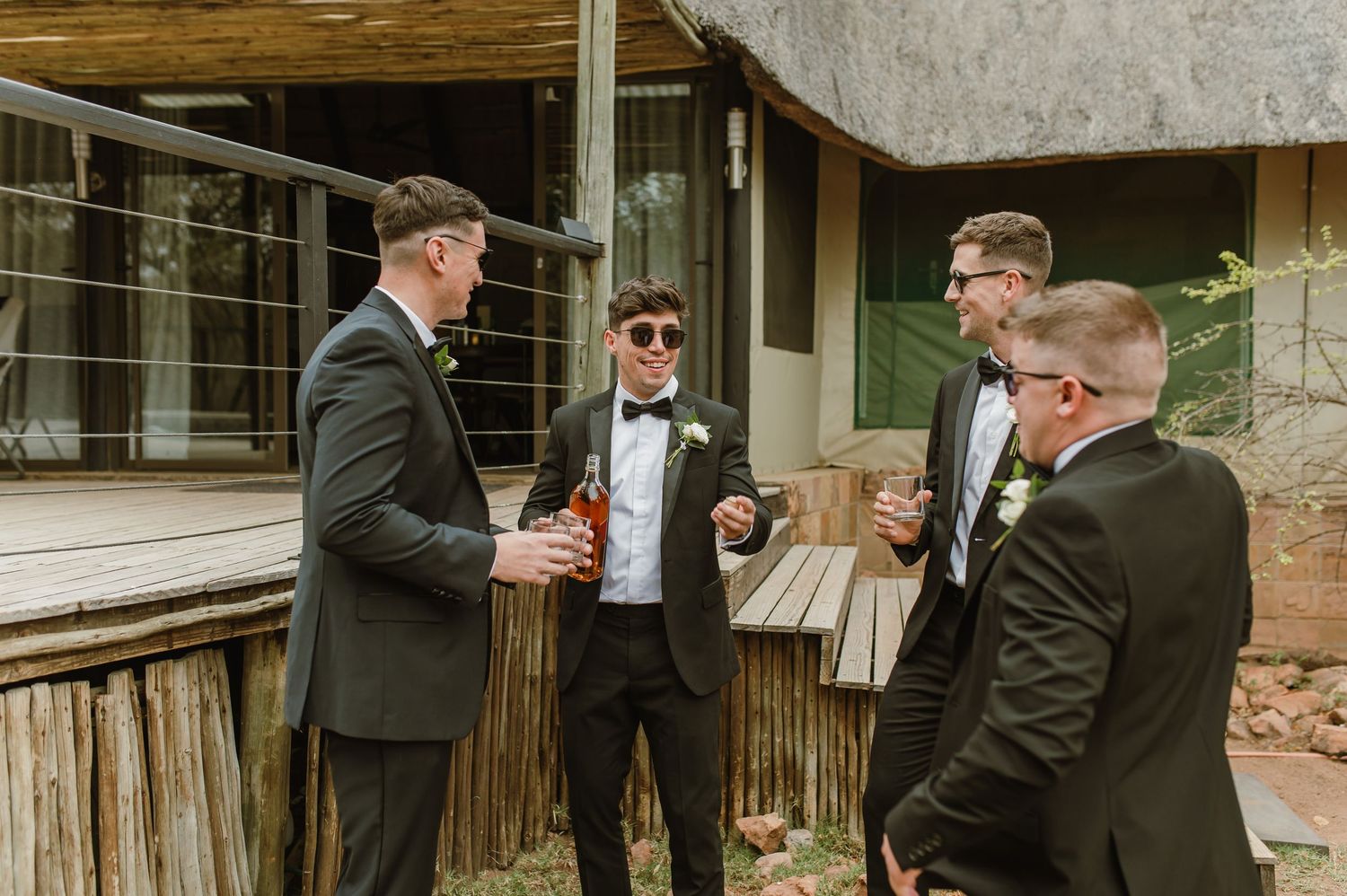 Groomsmen in black tuxedos and sunglasses share drinks and laughs on a rustic wooden deck.