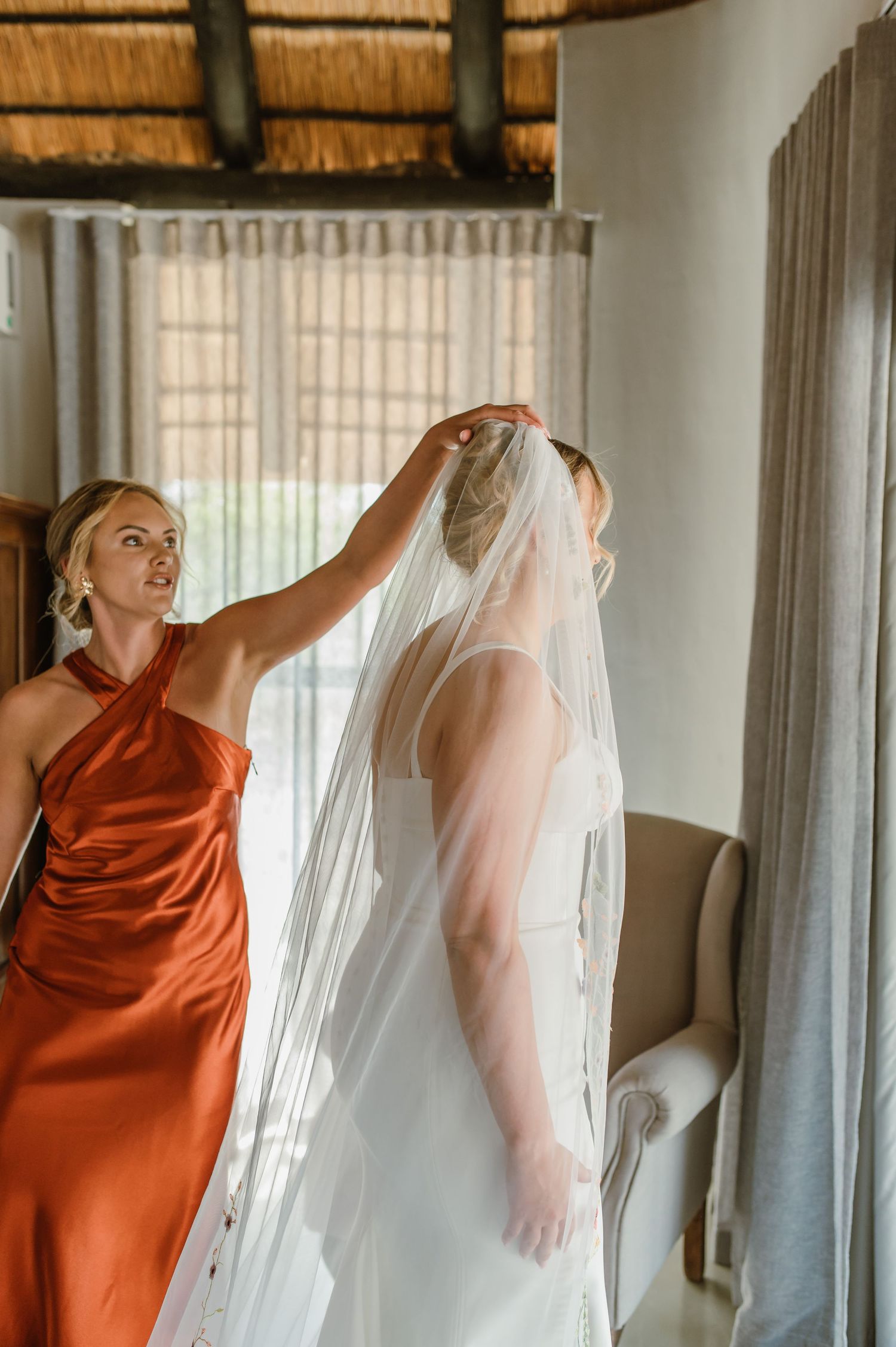 Bridesmaid in rust-colored dress helps adjust wedding veil in sunlit room.