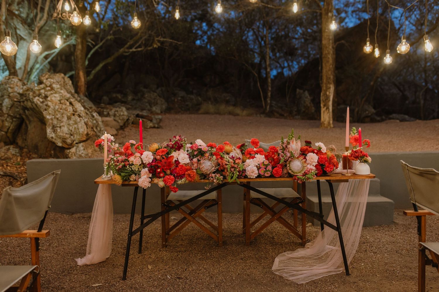 Elegant table settings with red flowers and candles under outdoor string lighting.