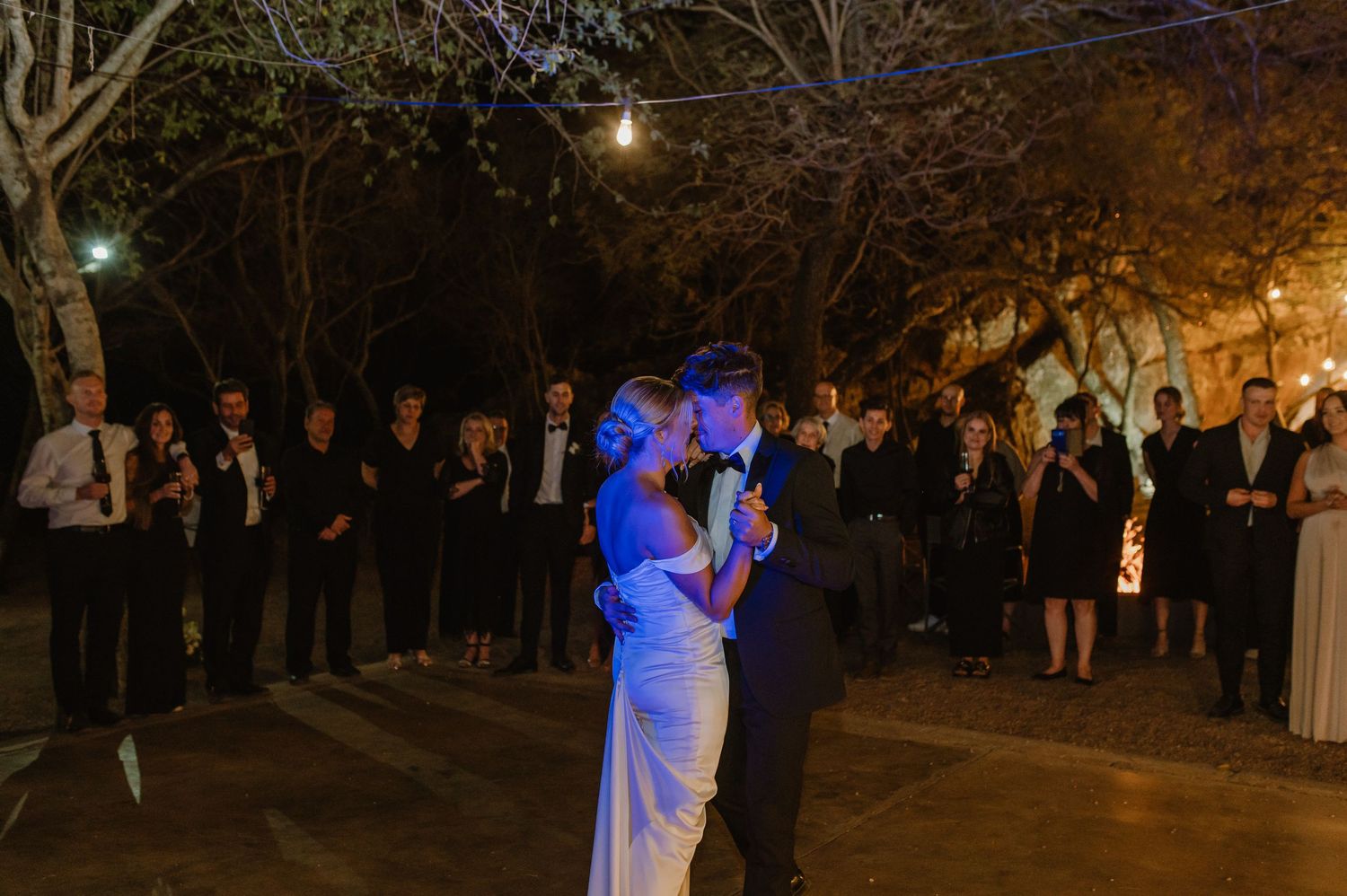 Evening wedding celebration with guests dancing under string lights and blue uplighting.