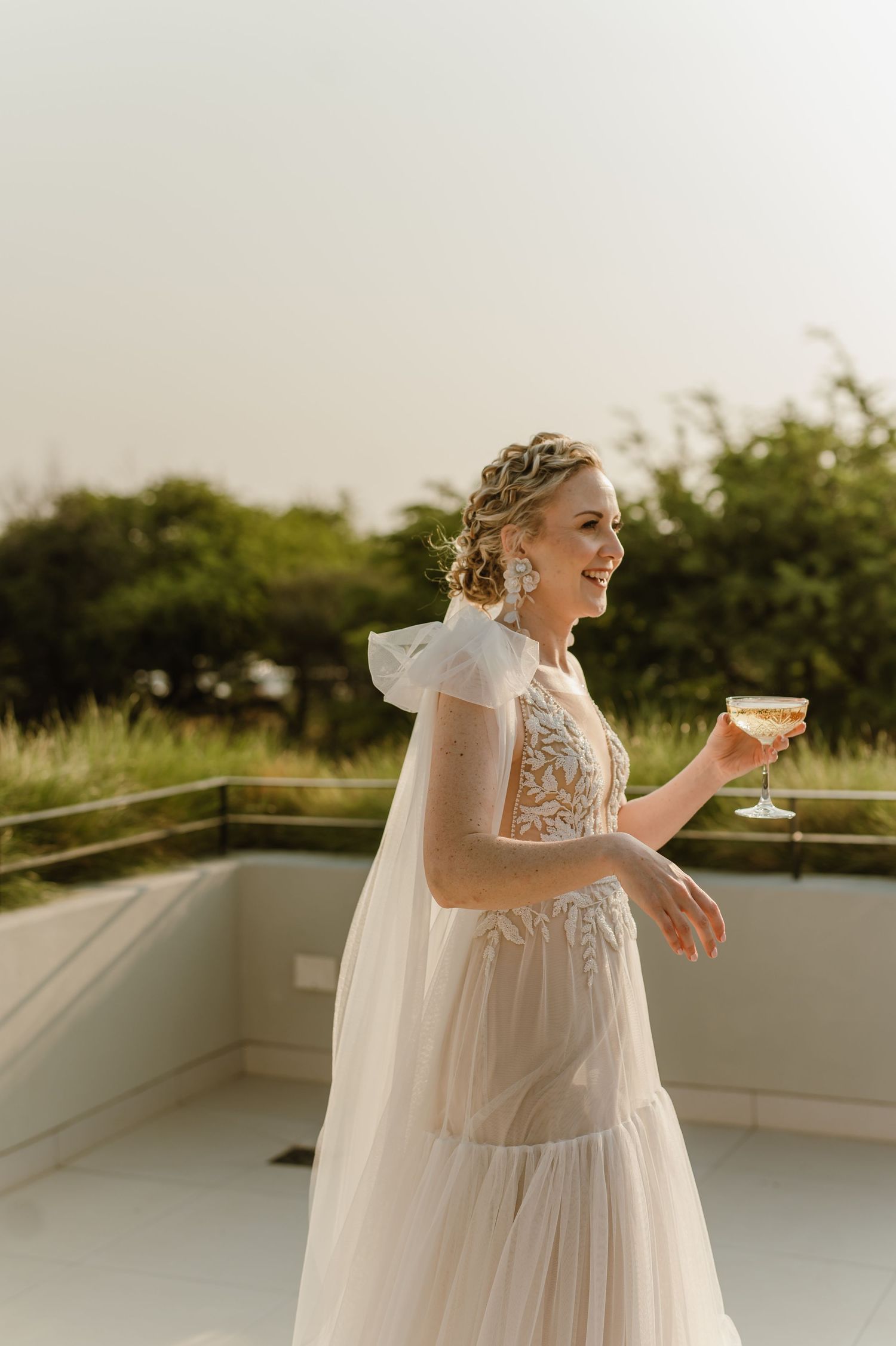 Bride in flowing white dress enjoying champagne on an outdoor terrace.