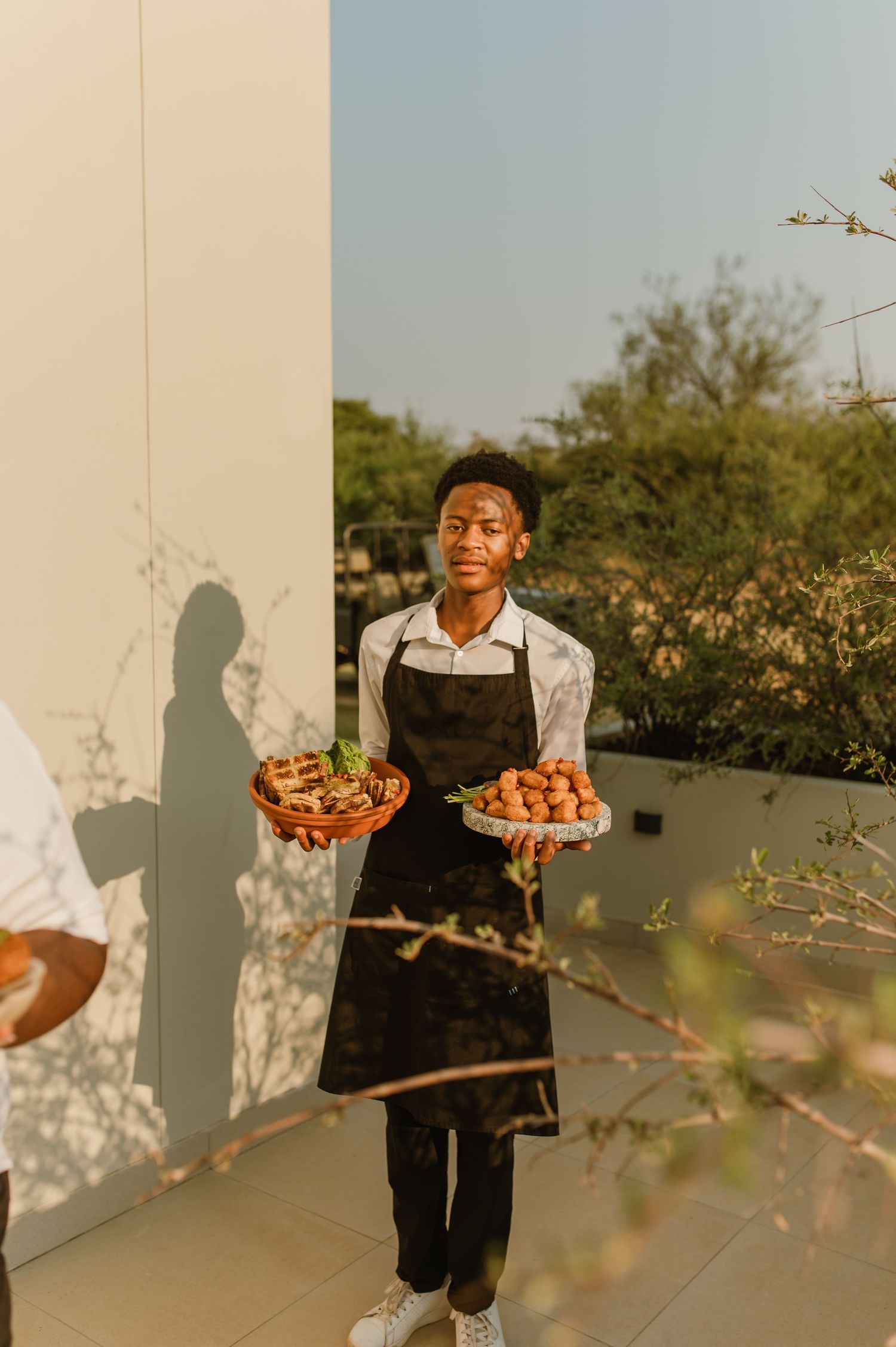 Server in black apron holds two platters of appetizers during an outdoor catering event at sunset.
