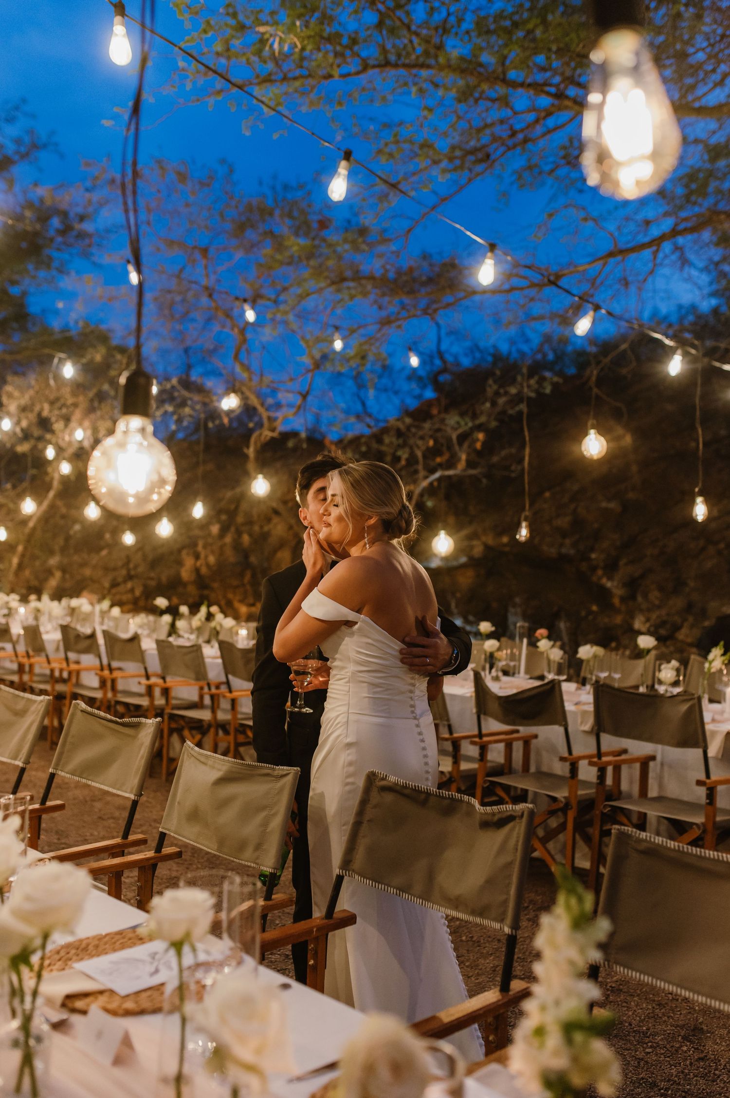 Outdoor wedding reception setup under string lights at dusk with elegant table settings and natural decor.