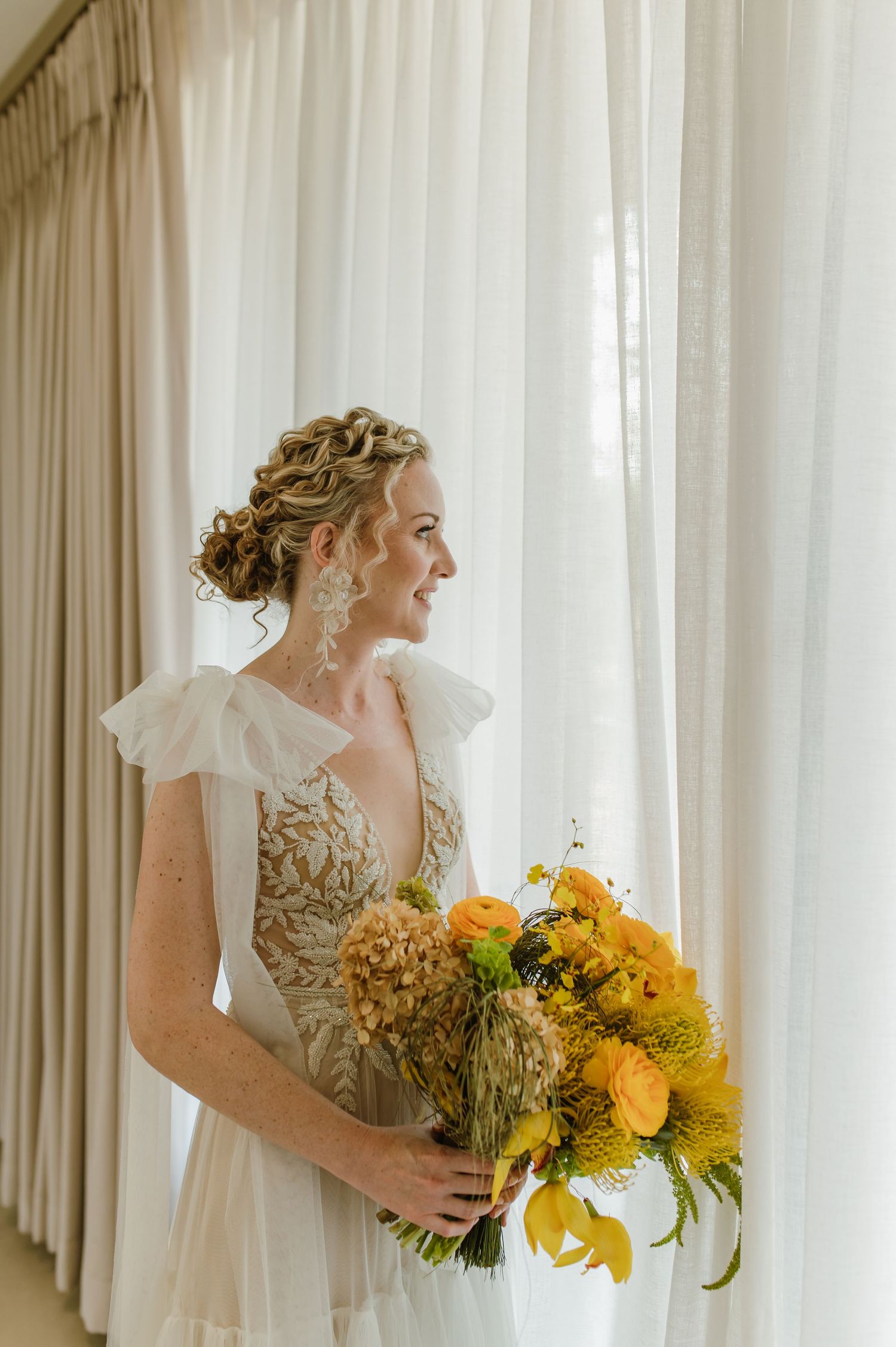 Bride holding vibrant yellow sunflower bouquet against white curtained window.