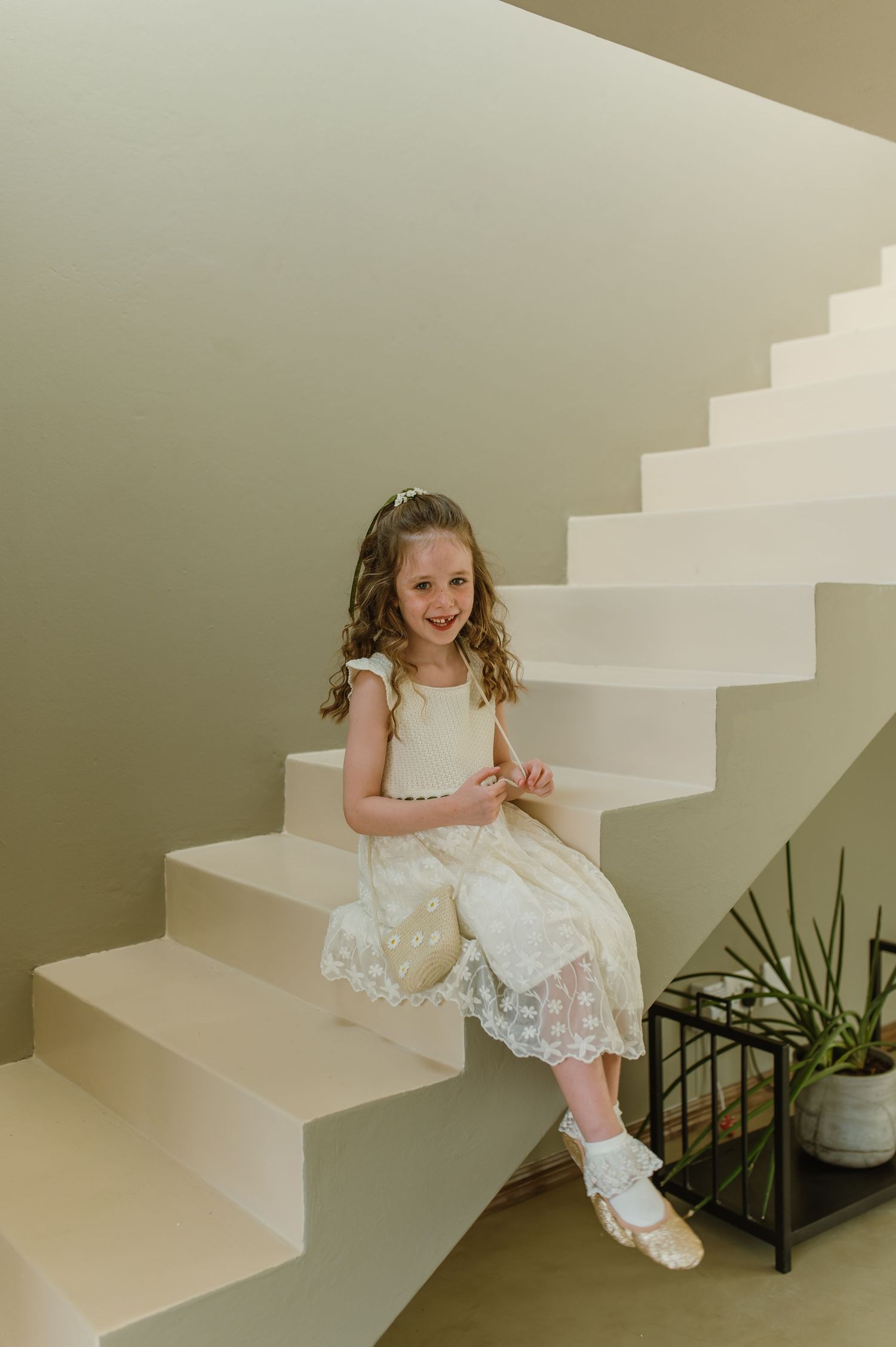 A young flower girl in a white lace dress sits playfully on white stairs.