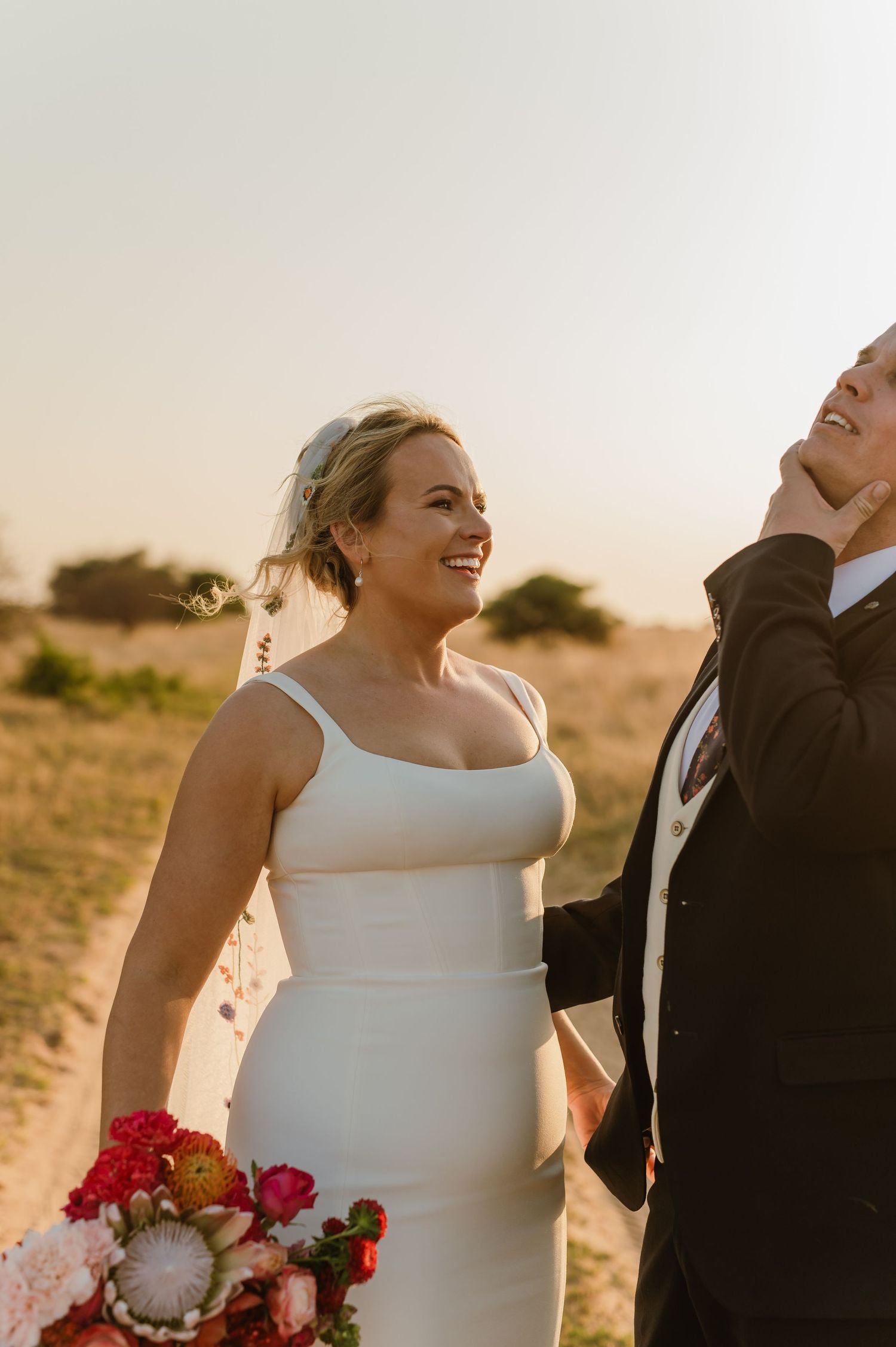 A joyful wedding couple laughs together in warm evening light holding vibrant red flowers.