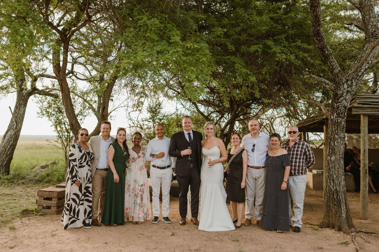 Group photo of wedding party and guests lined up under trees at outdoor venue.