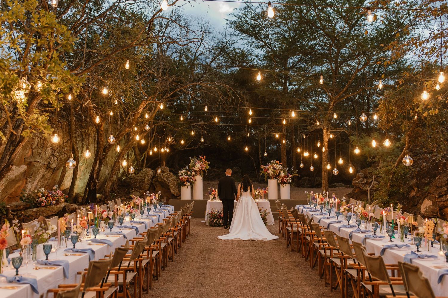 Elegant outdoor wedding reception under string lights with long tables set among trees at dusk.