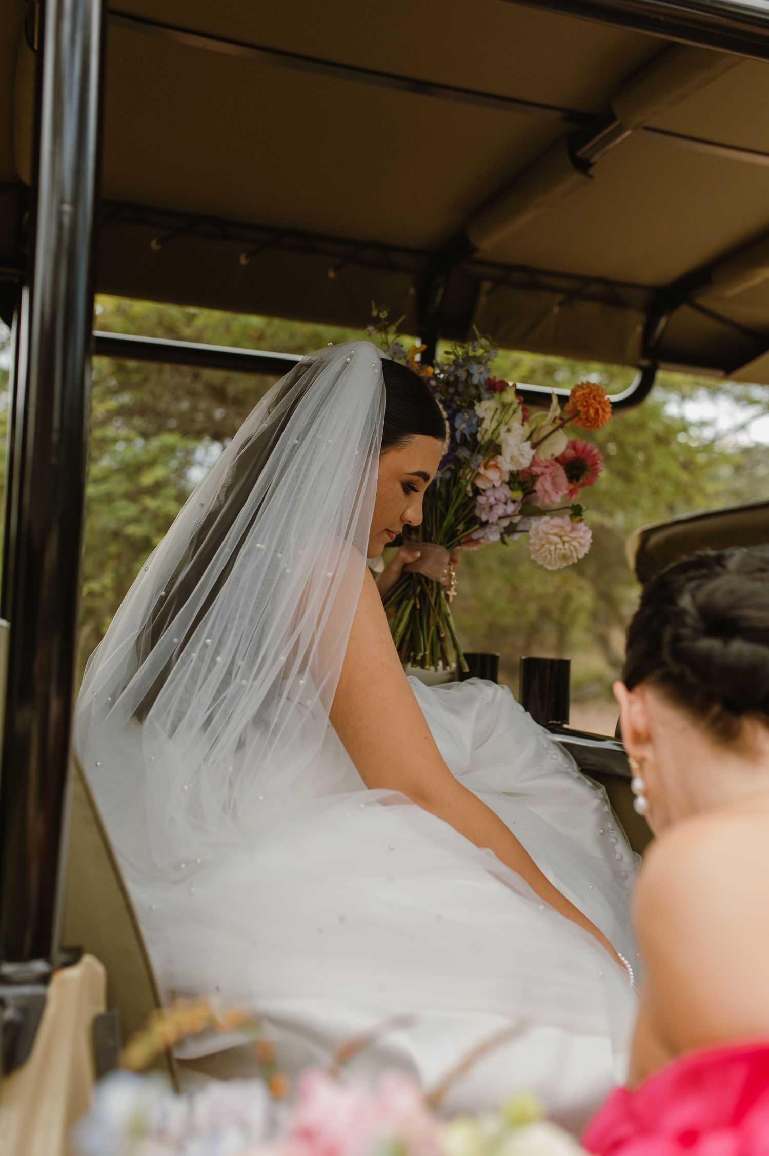 Bride in white gown and veil holding colorful bouquet while seated in a vintage wedding vehicle.