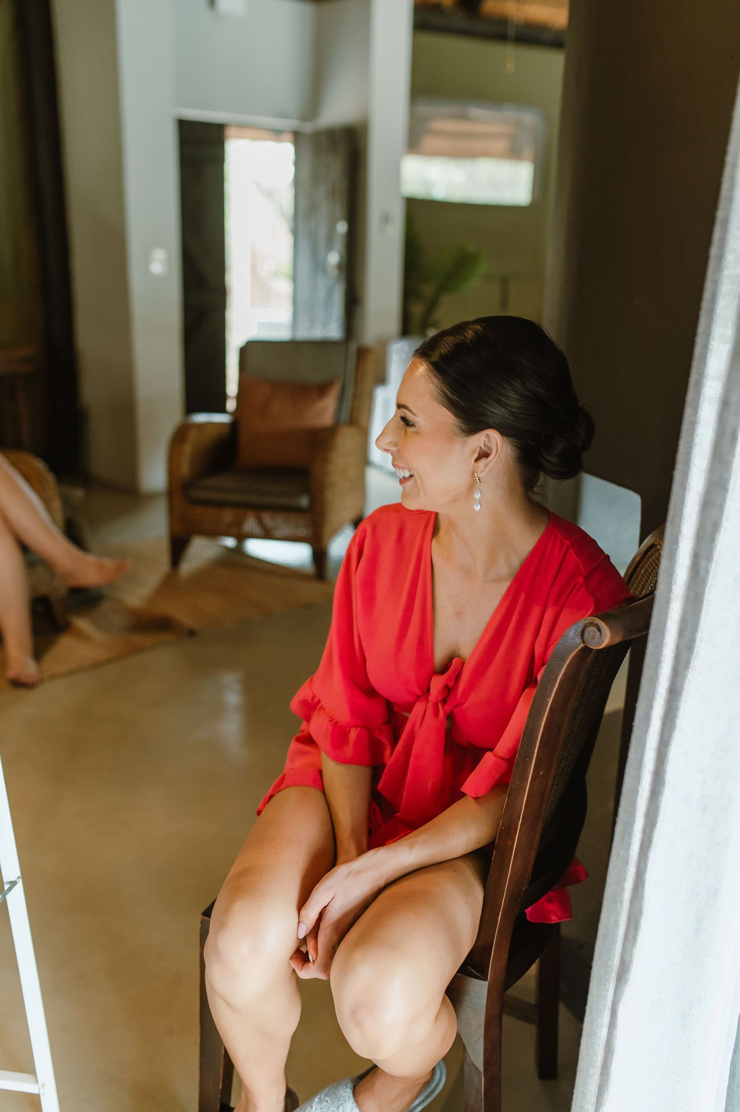Smiling bridesmaid in red dress sitting on wooden chair during wedding preparations indoors.