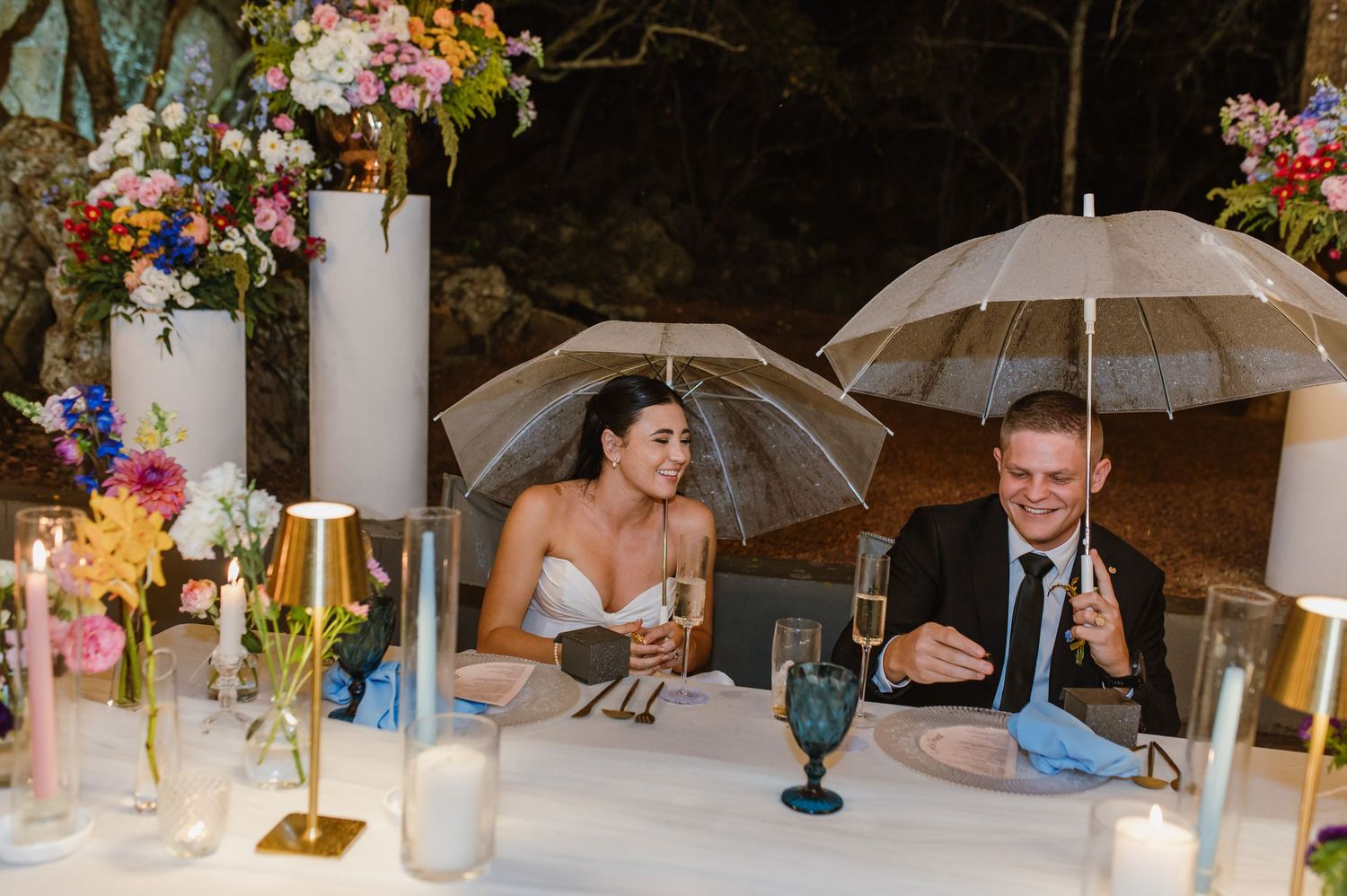 Bride and groom laugh at sweetheart table holding champagne and clear umbrellas during rainy reception.