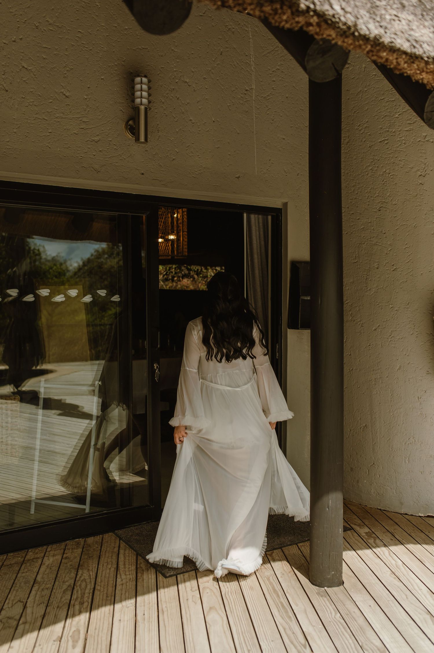 Bride in a sheer white robe entering a modern safari lodge through glass sliding doors on a wooden deck.
