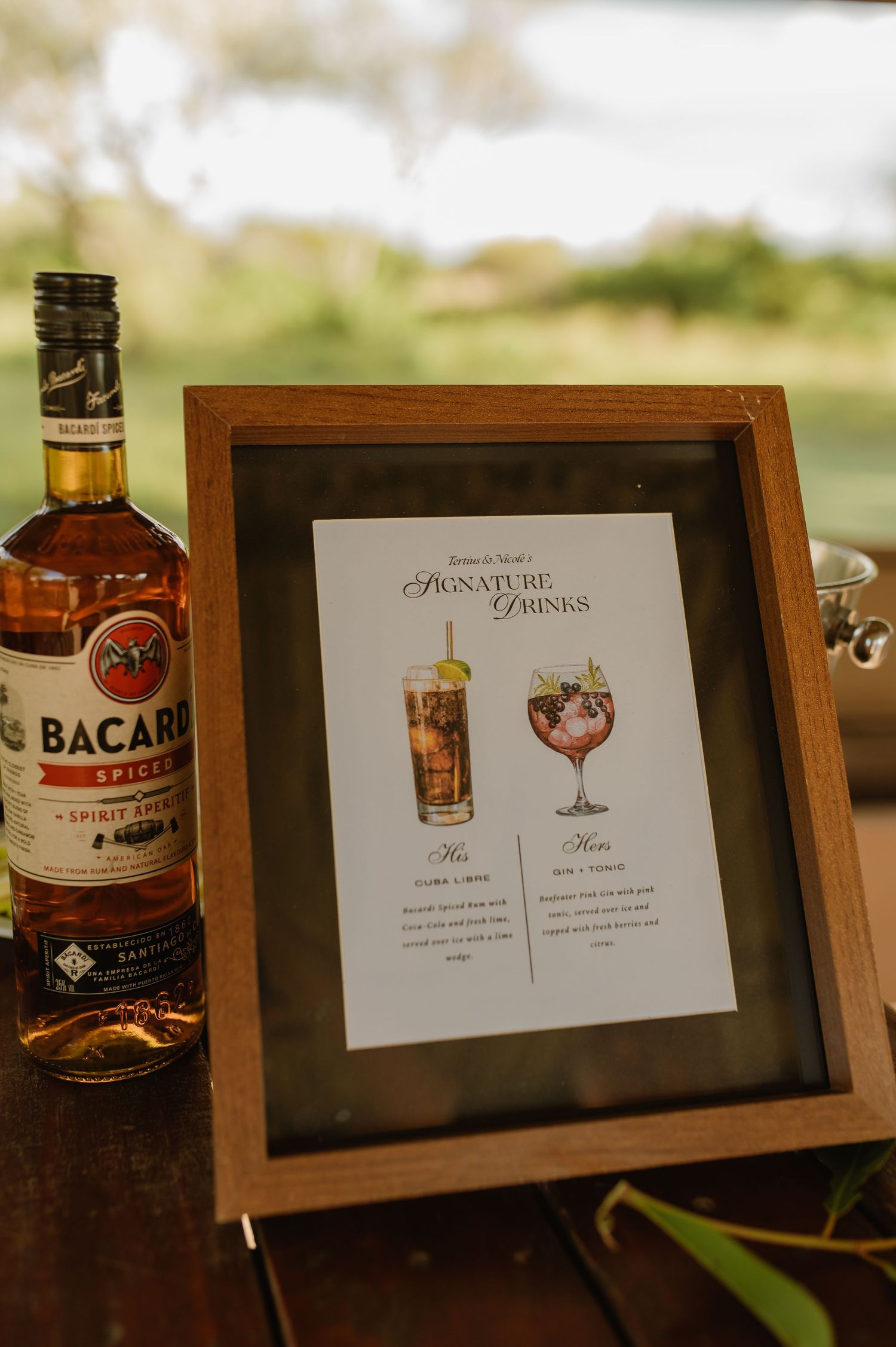 Framed signature drinks menu beside Bacardi Spiced rum bottle at outdoor wedding reception bar.