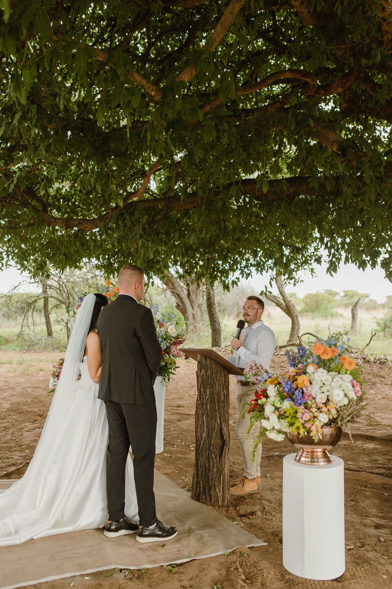 Bride and groom exchange vows under a lush tree at an outdoor wedding ceremony with colorful florals.