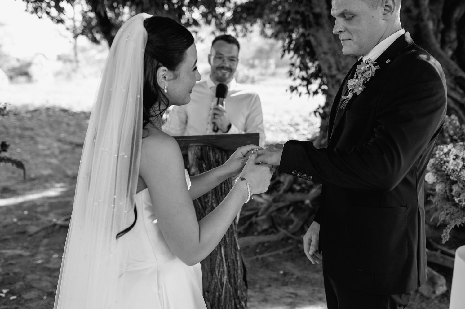Black and white photo of bride placing ring on groom's finger during outdoor wedding ceremony.