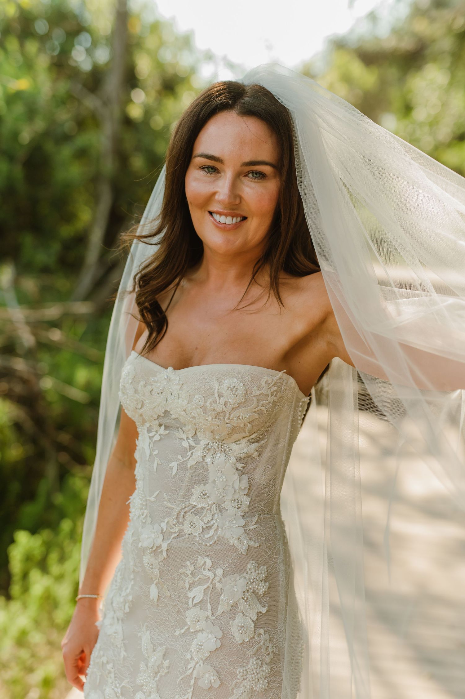 A smiling bride in a strapless lace gown with a flowing veil in a sunlit outdoor setting.