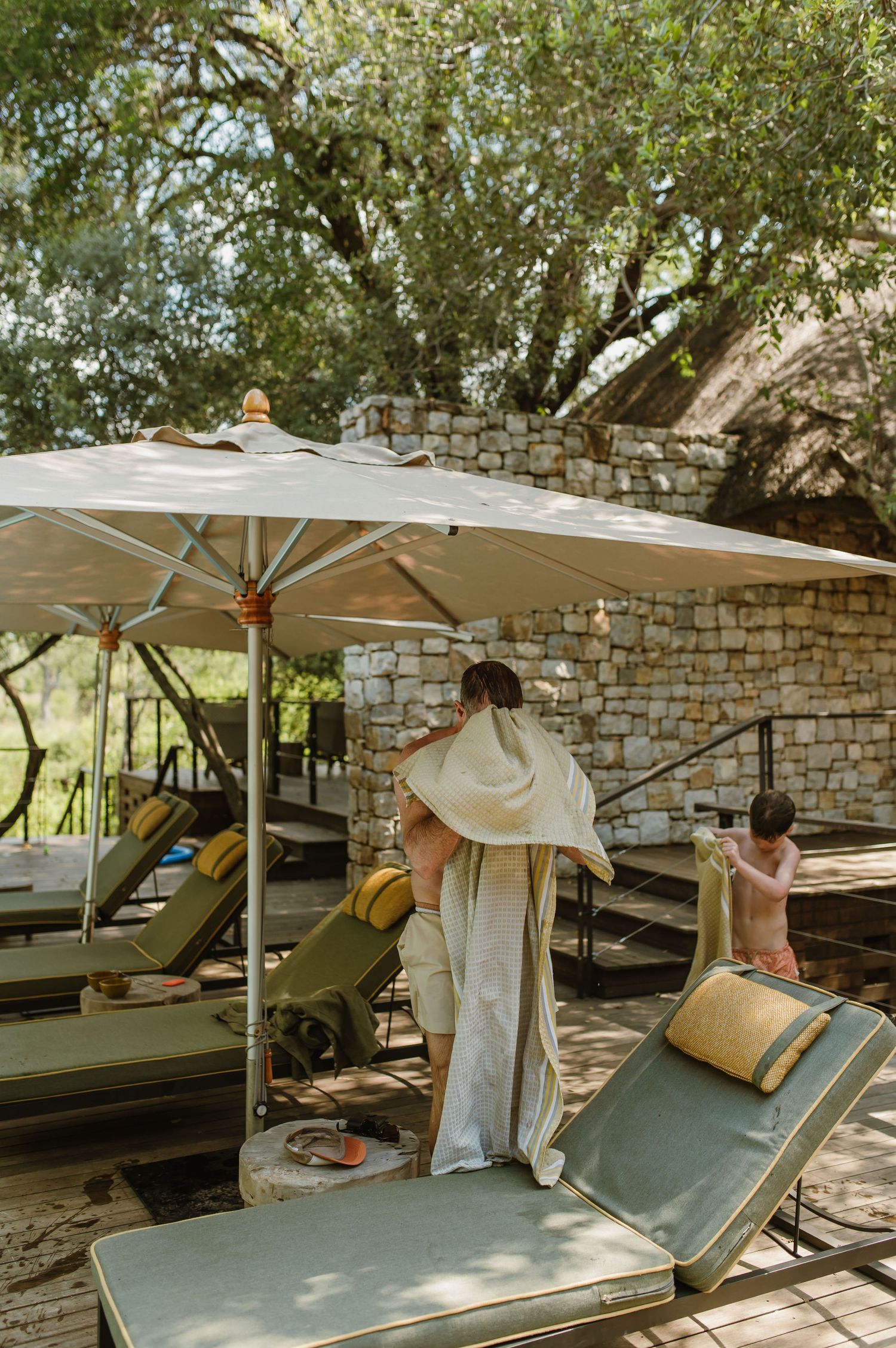 A woman wrapped in a white towel stands near lounge chairs under a large umbrella at a safari lodge.