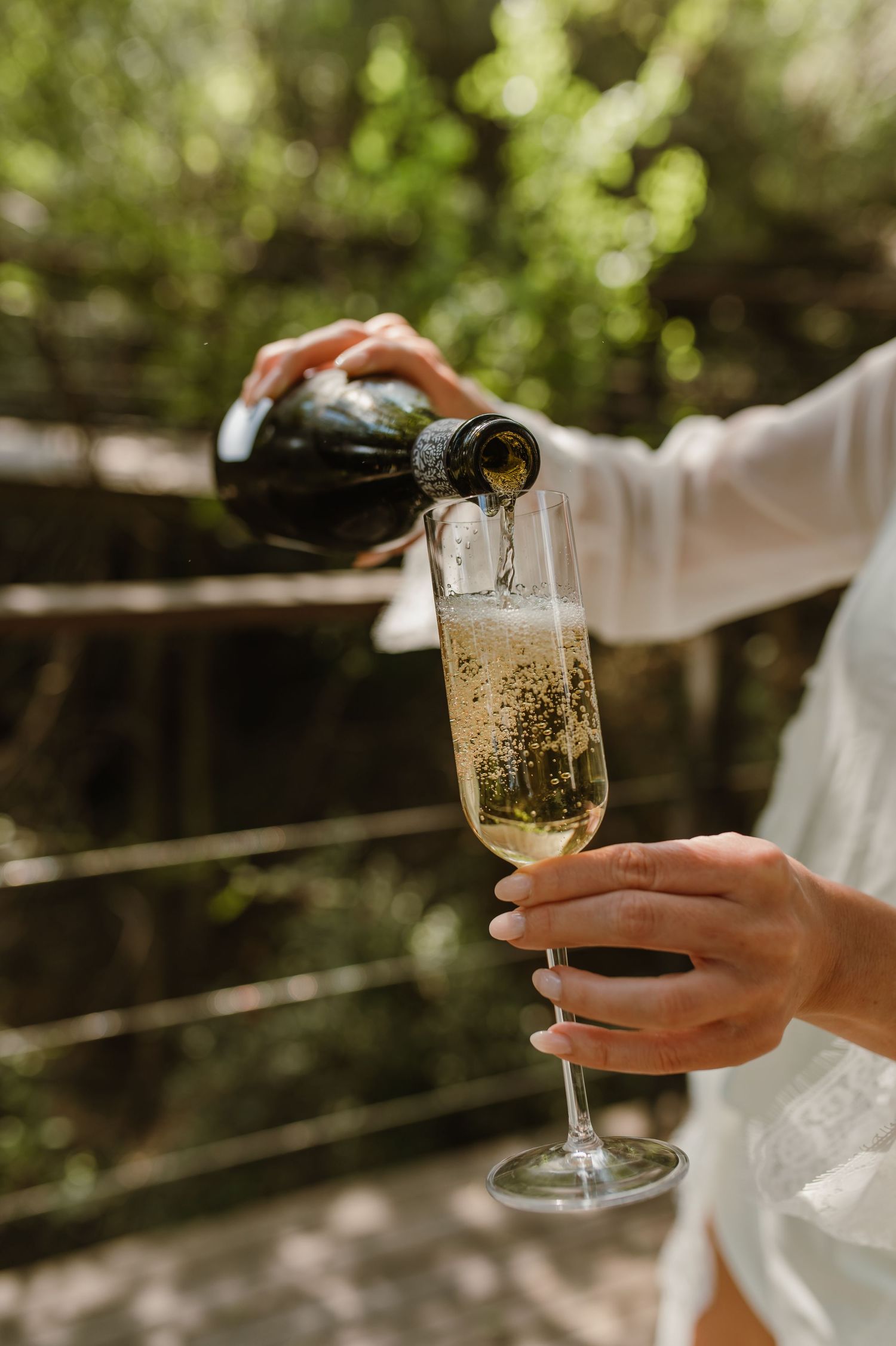Person in white robe pouring sparkling champagne into a flute glass outdoors with green foliage.