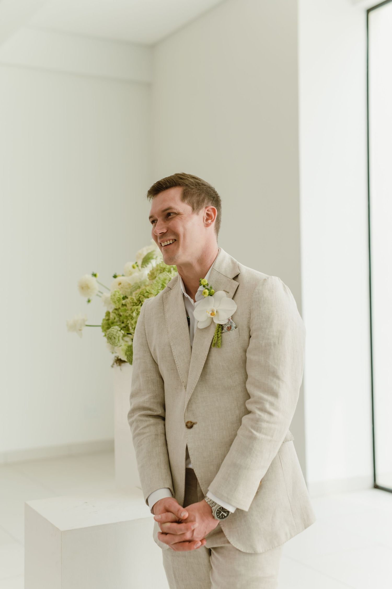 Wedding attendee in a light beige suit and boutonniere standing in a bright modern space.