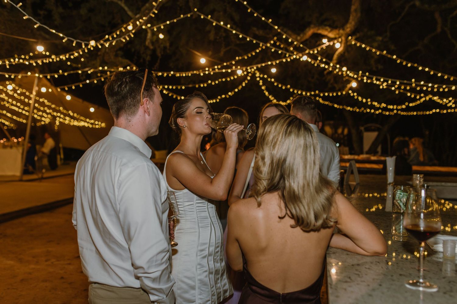 Wedding celebration under string lights with guests enjoying drinks and conversation.