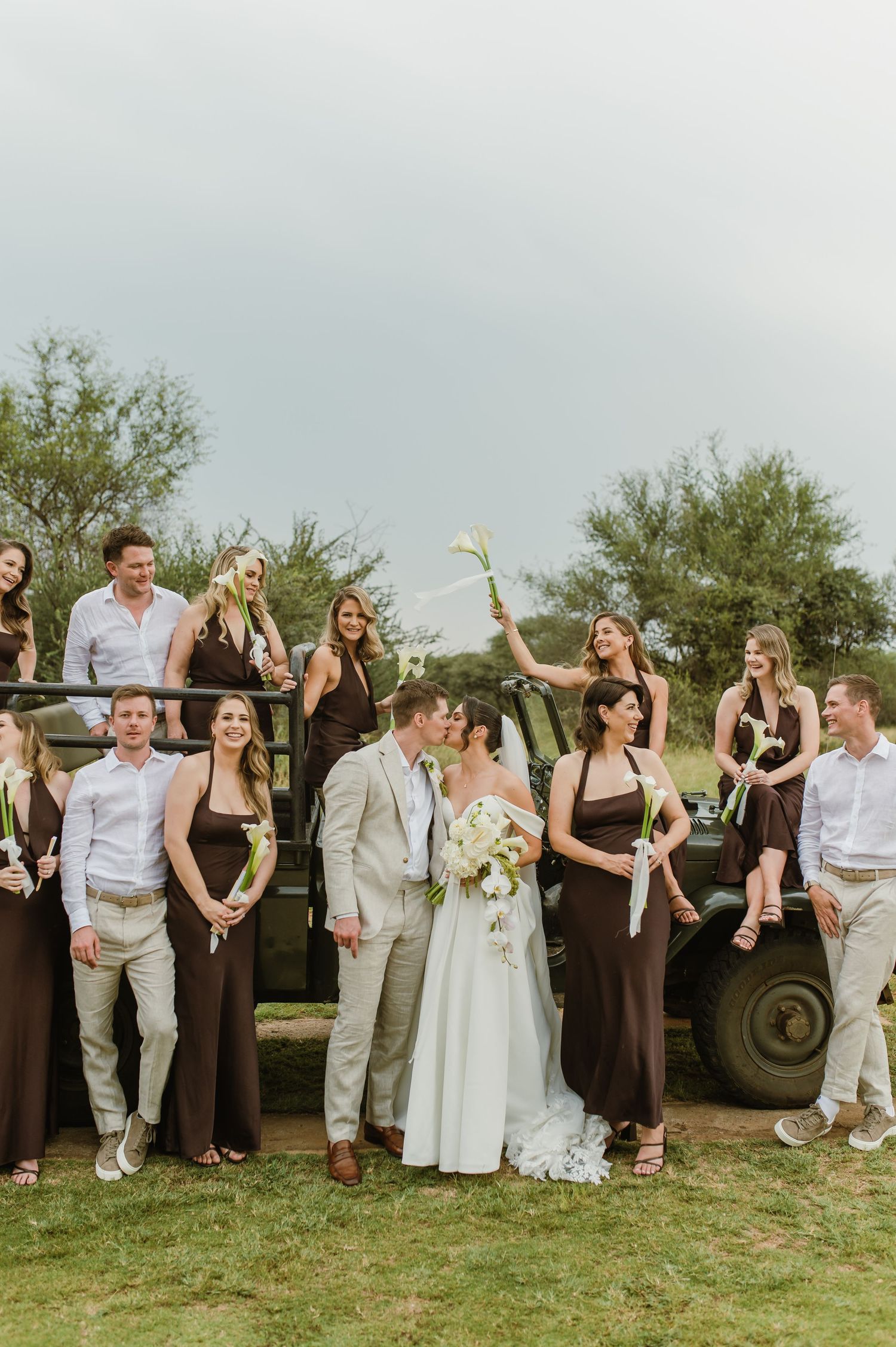 Wedding party poses together outdoors with bridesmaids in black dresses and groomsmen in light suits next to rustic decor.