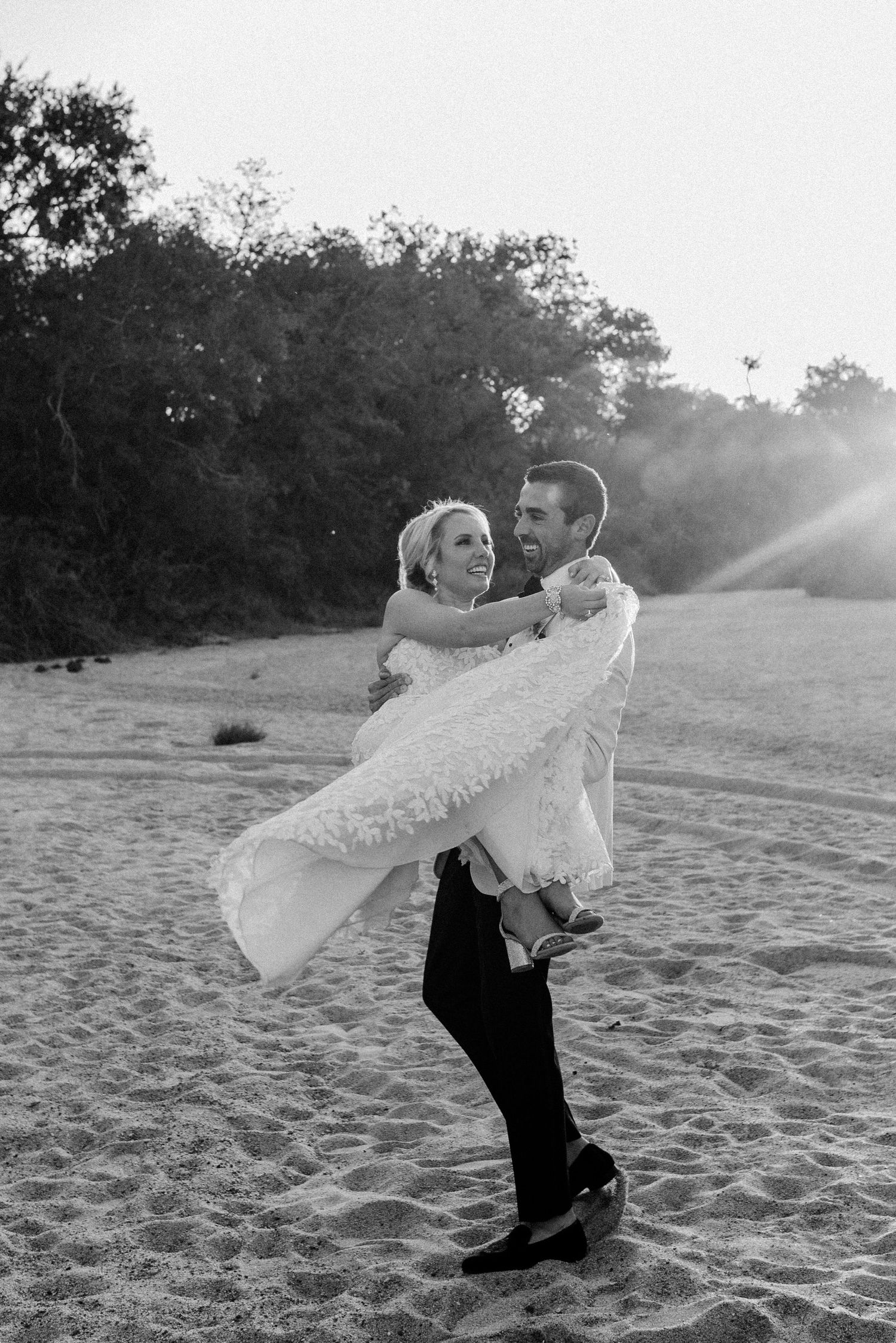 Black and white wedding photo of couple embracing on beach at sunset.