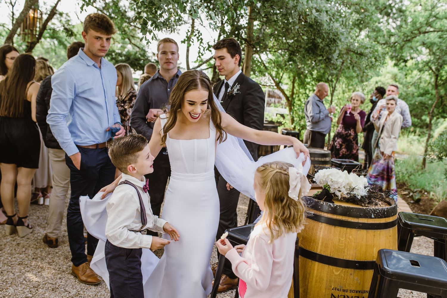 Wedding celebration with children and guests gathered around wooden barrels in an outdoor garden setting.