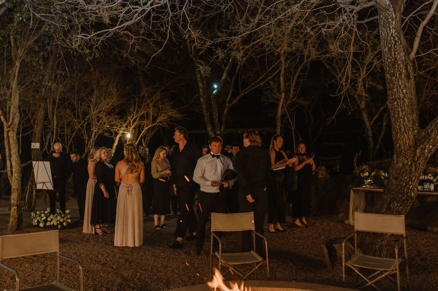 Guests mingling at an outdoor evening event under natural tree canopy.