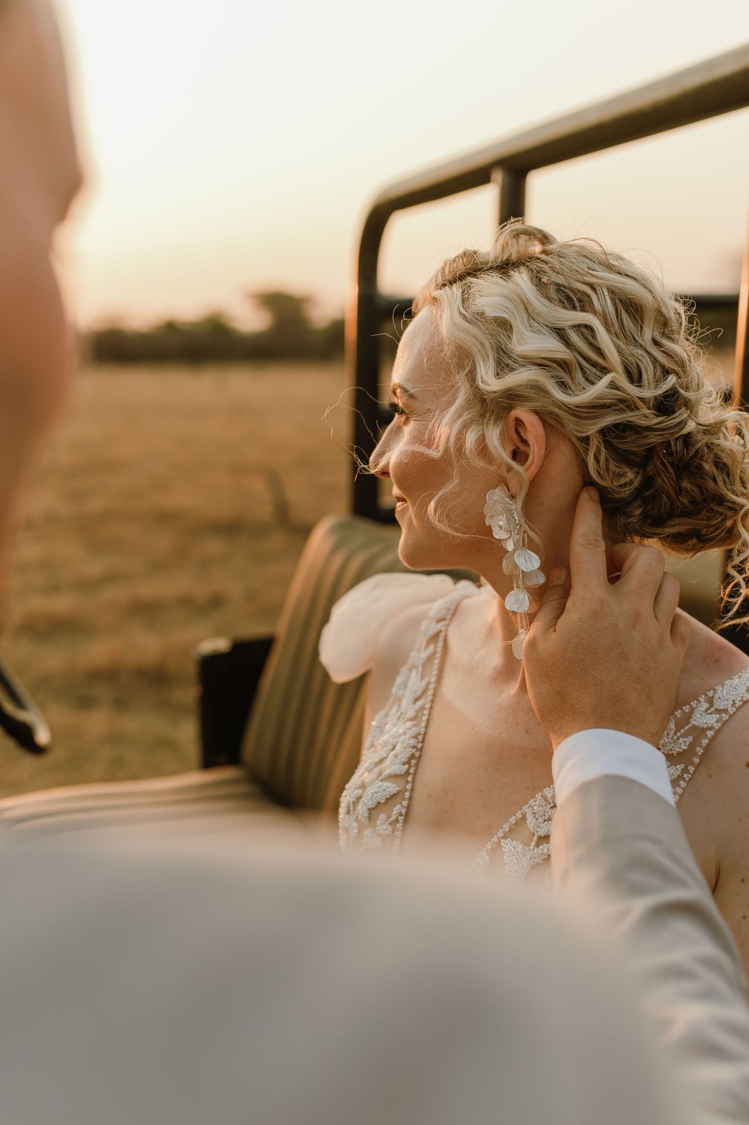 Candid moment of a bride in an elegant lace gown sitting in a safari vehicle at sunset.