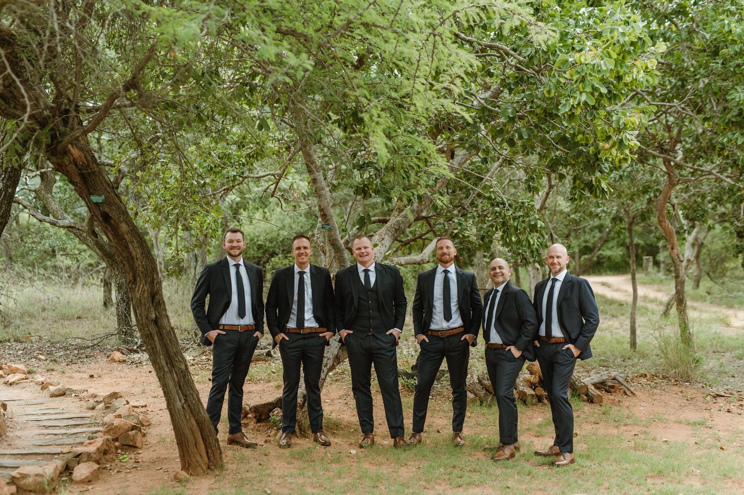 Wedding party in black suits lined up along trees in an outdoor garden setting.