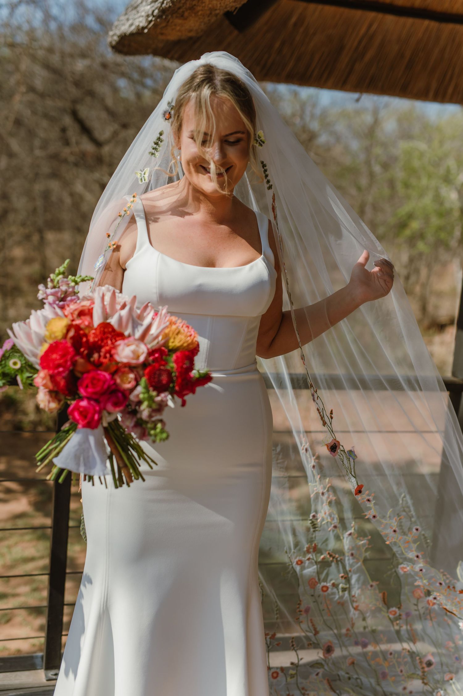 Bride in elegant white dress holds a vibrant red and pink bouquet under a thatched pavilion.