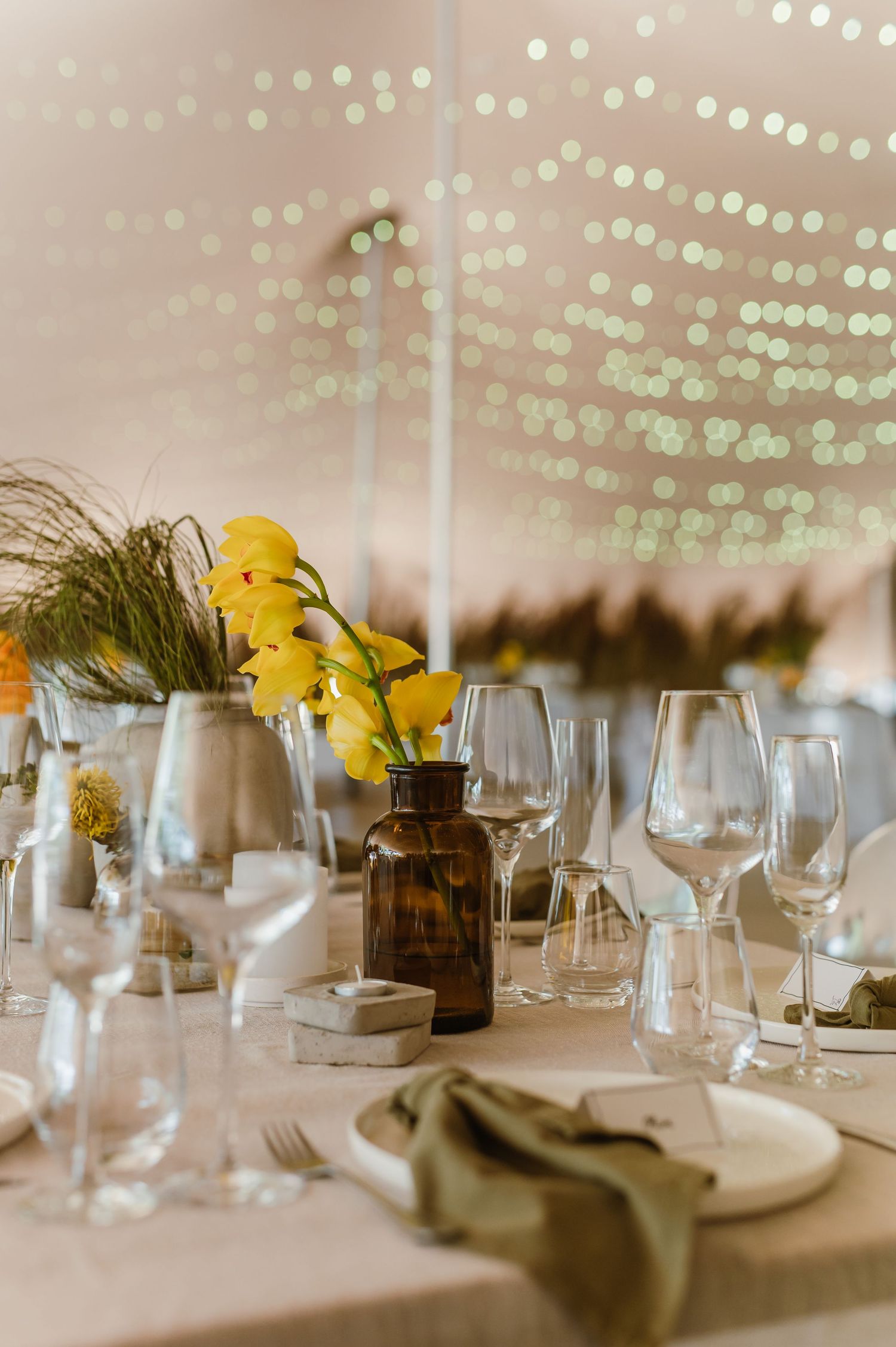 Wedding table settings with wine glasses and yellow flower centerpieces under string lights.