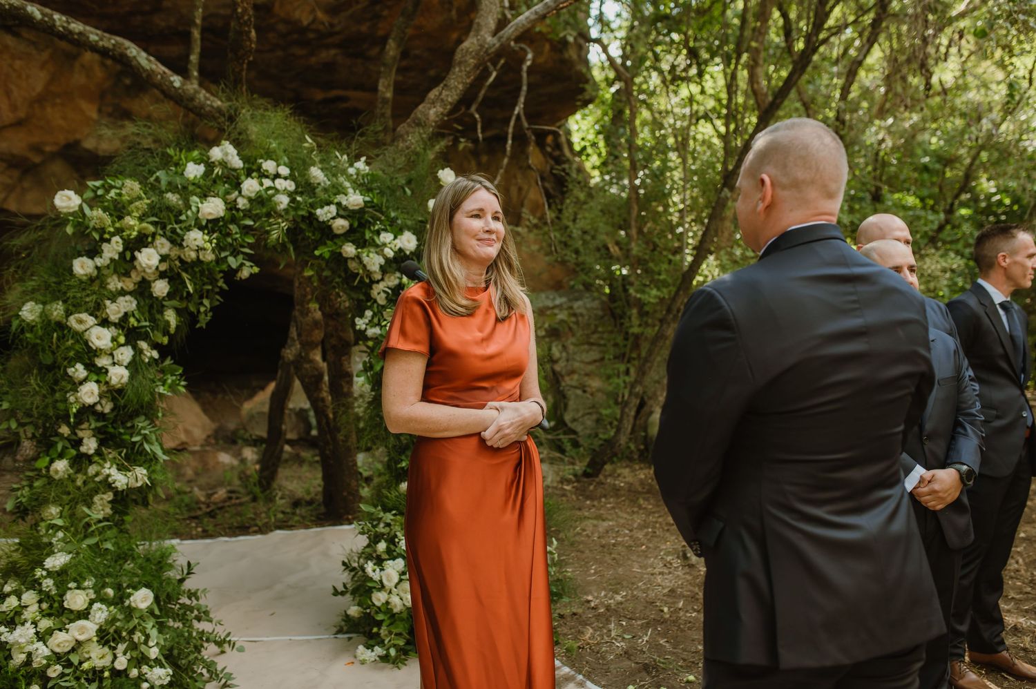 Wedding guests gather in a natural wooded setting with flowering bushes and trees.