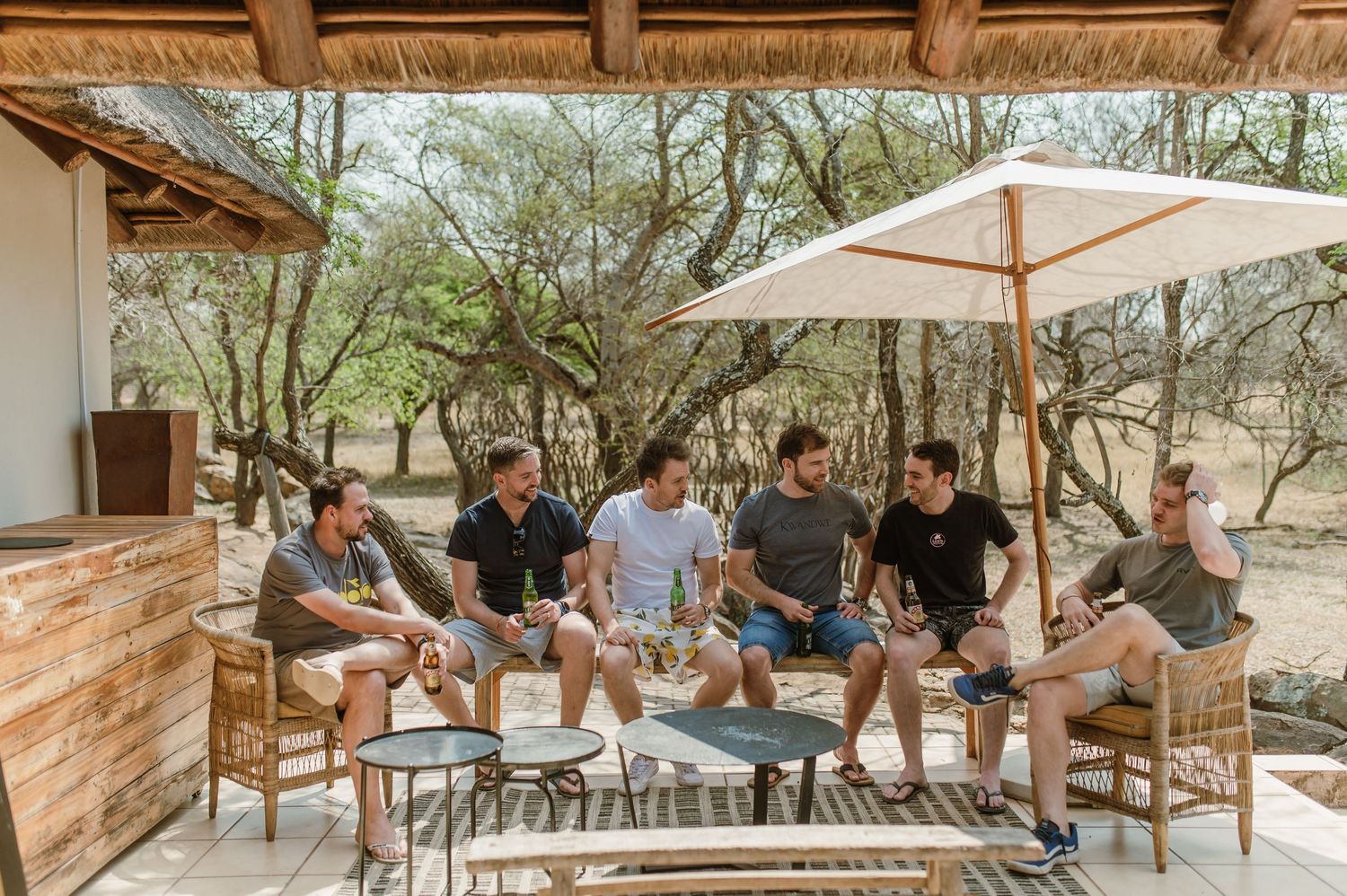 A group of people relaxing under a wooden pergola with umbrellas in an outdoor setting.