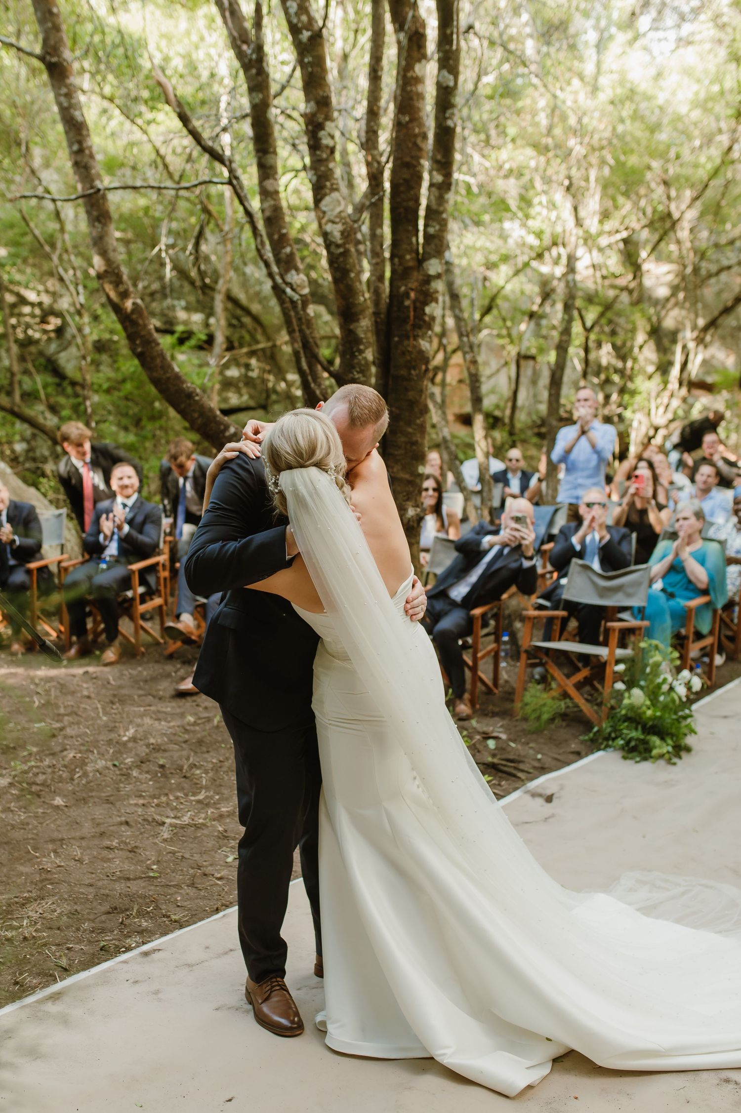 Couple celebrates during their woodland wedding ceremony as guests look on from wooden benches.