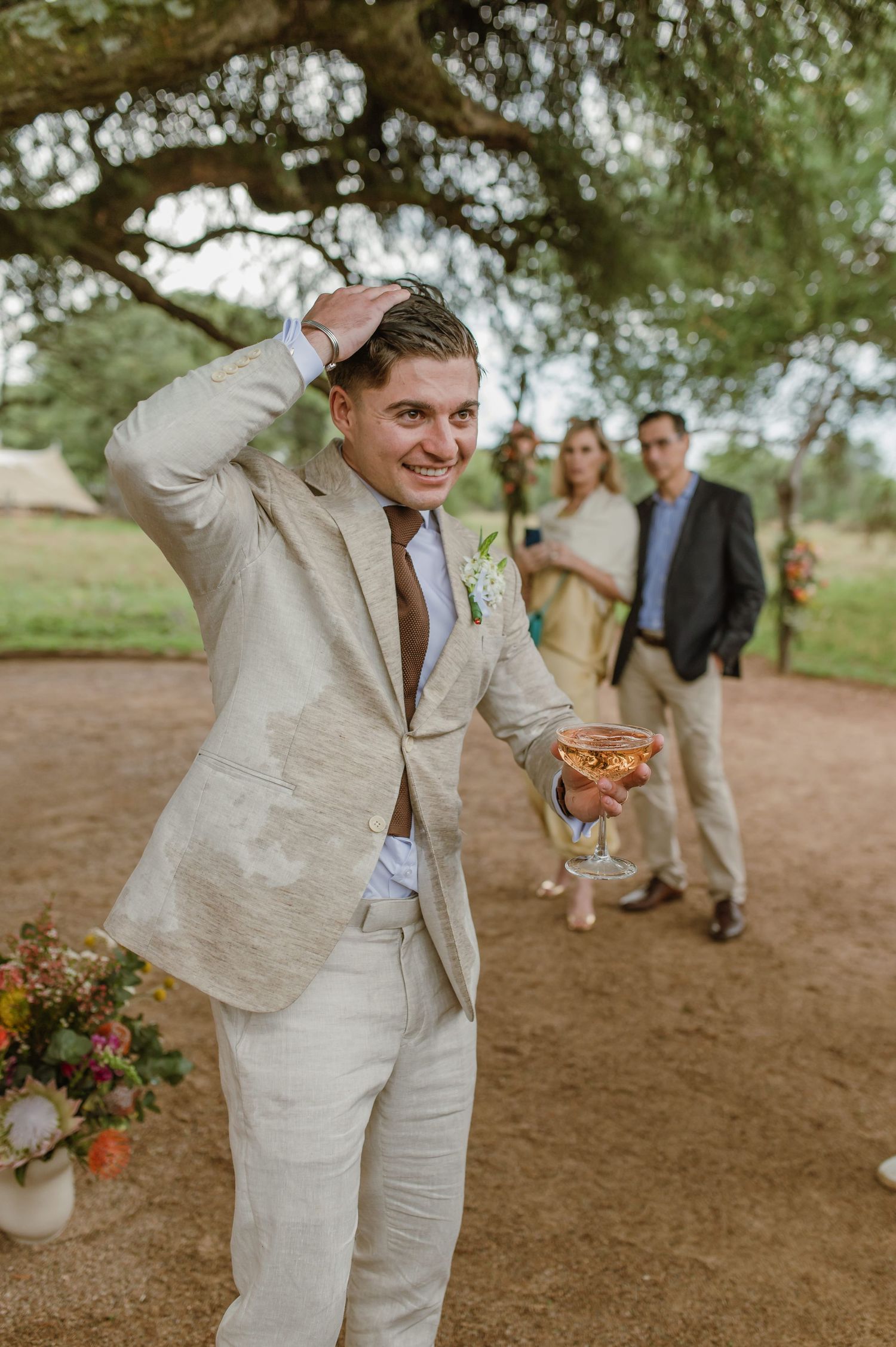 Wedding celebration under a tree with guests visible in background and champagne toasts.