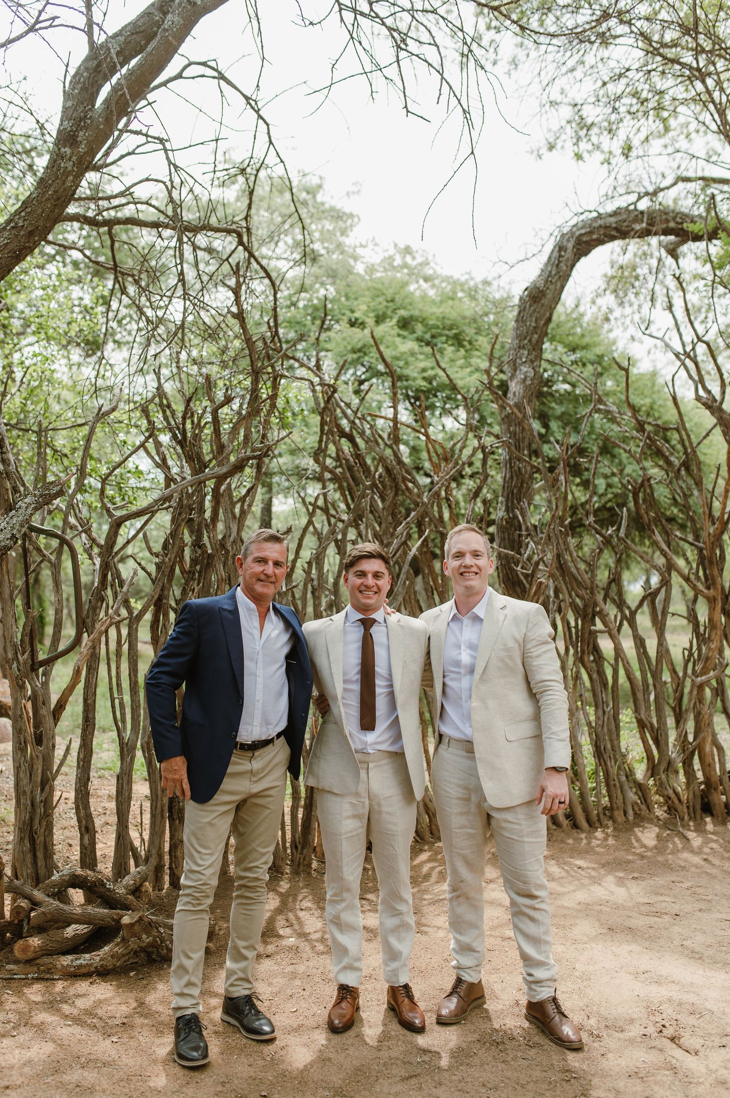 Wedding party members pose casually on a dirt path surrounded by trees.
