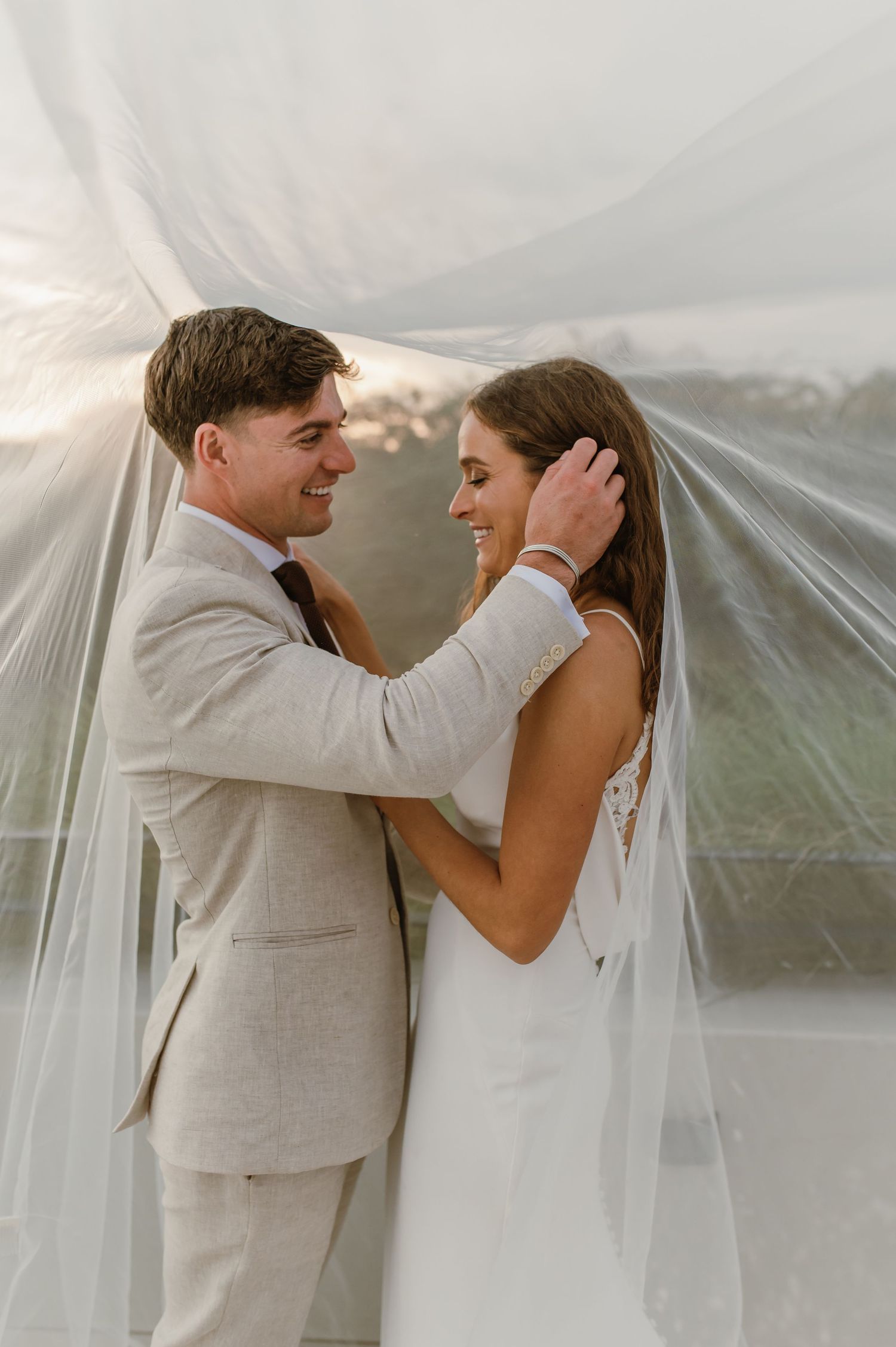 Joyful wedding couple shares loving moments beneath flowing veil at outdoor venue.