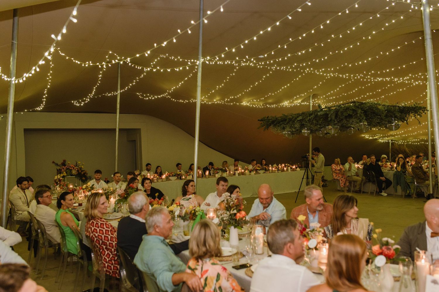 Wedding reception guests seated at long tables under a tent with twinkling string lights overhead.
