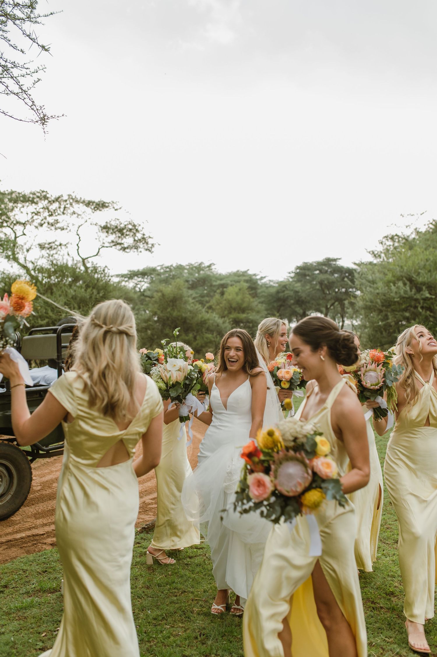Candid moment of wedding party in yellow dresses sharing laughs while holding bright floral arrangements.