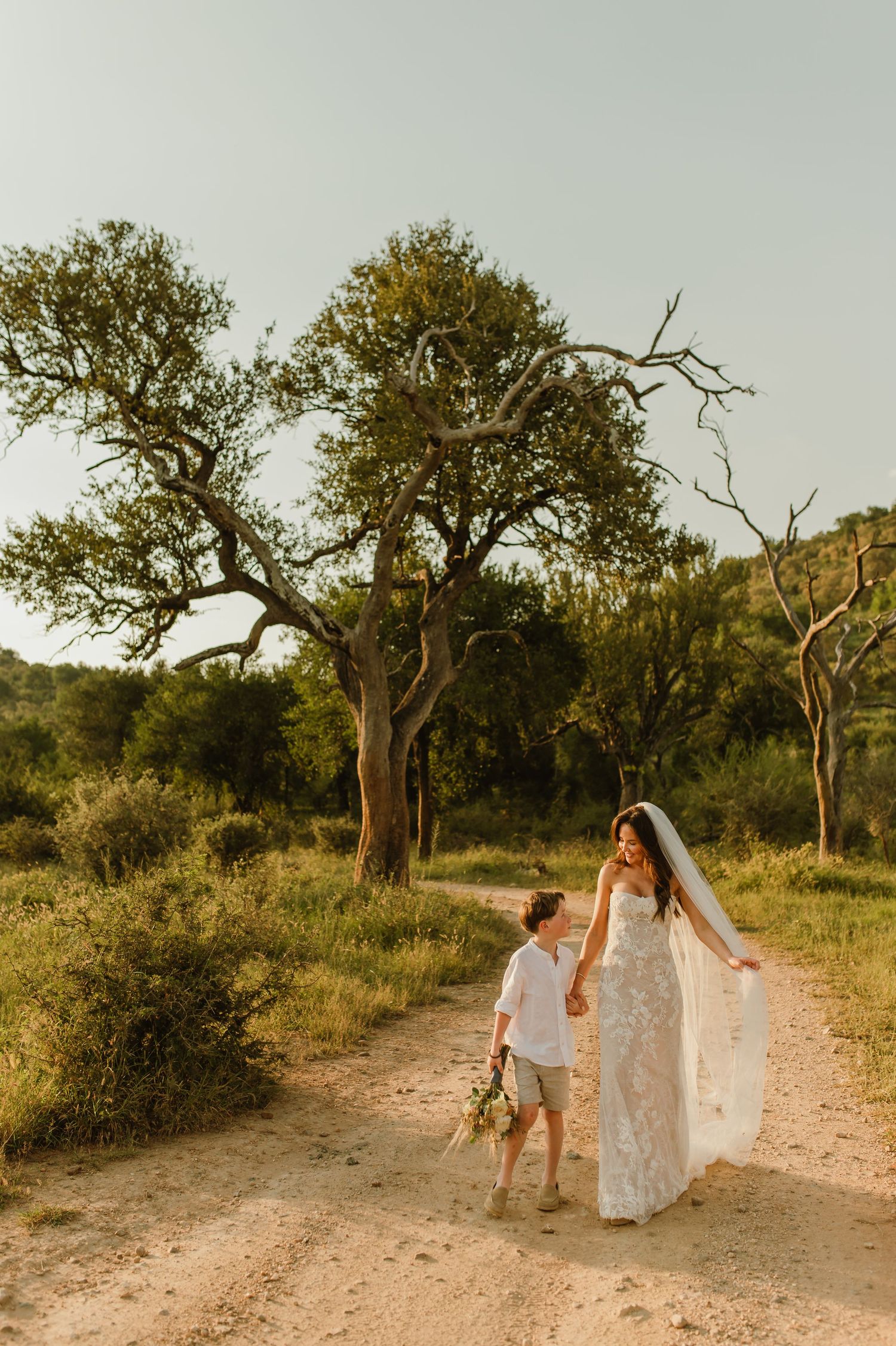 Bride in lace gown walks hand-in-hand with young boy on sandy path surrounded by lush trees at golden hour.