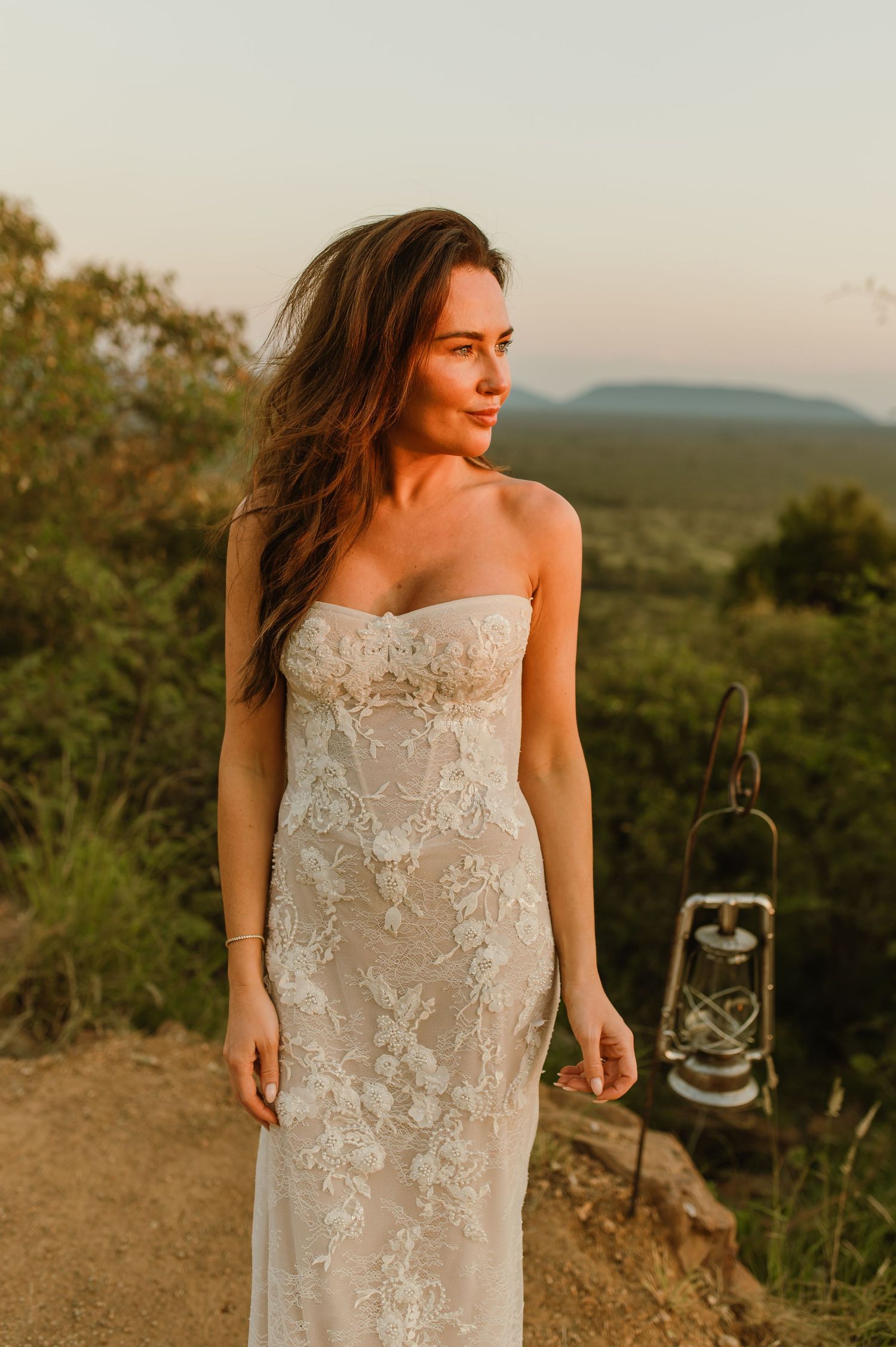 Bride in strapless floral lace gown stands at sunset with mountain backdrop and vintage lantern nearby.